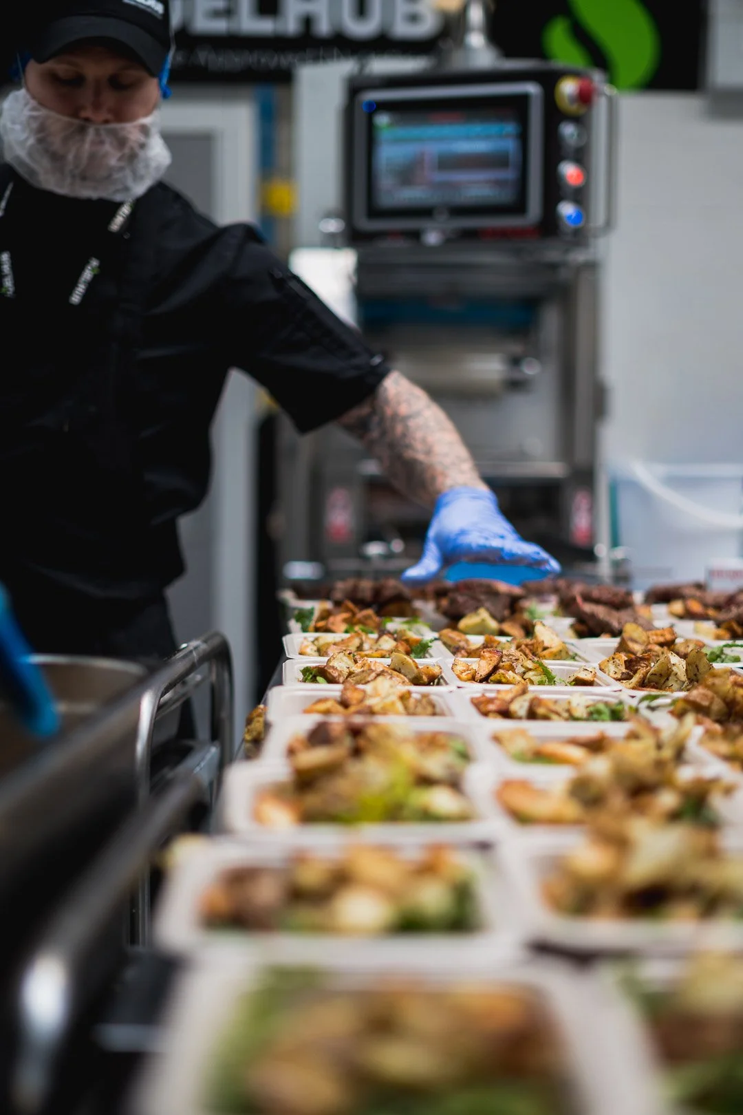 A chef wearing a black uniform, blue gloves, and a hairnet prepares multiple food trays with cooked vegetables and meat in a commercial kitchen kitchen.