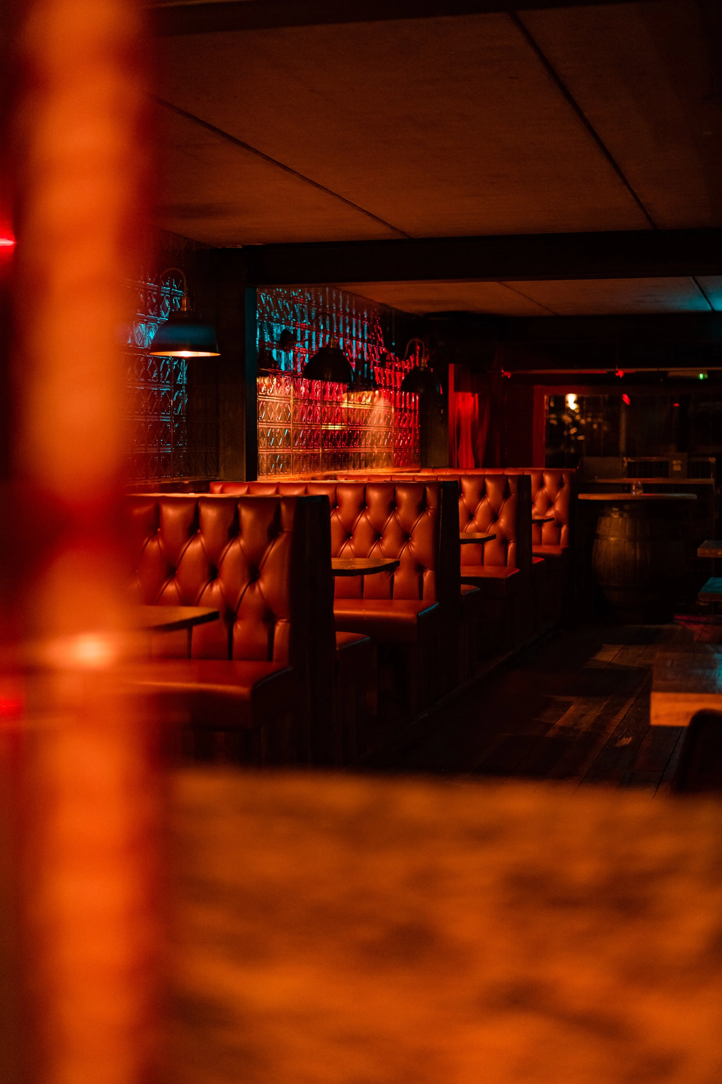 Dimly lit bar or lounge with tufted leather booths, dark wood furniture, and colorful neon lighting.