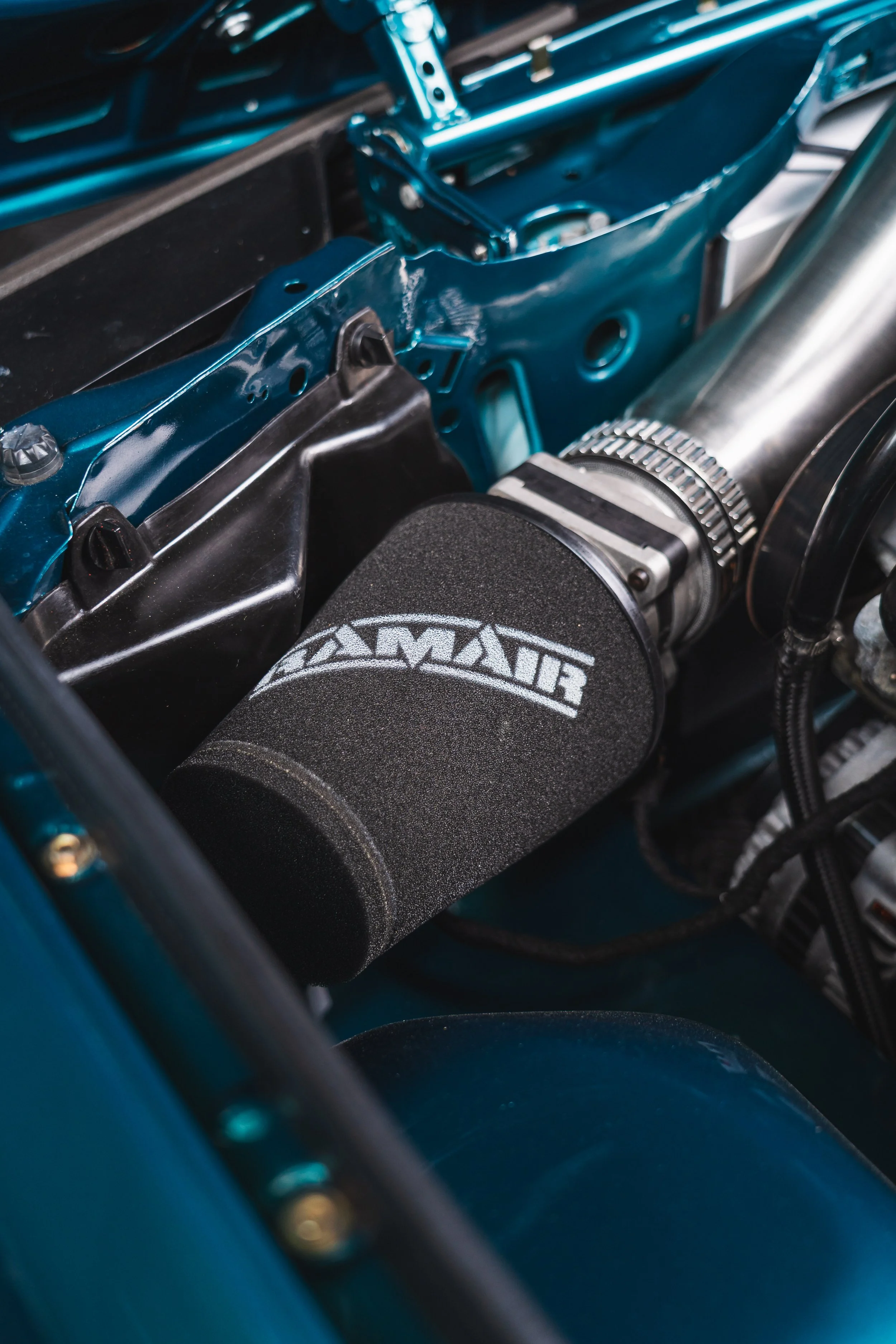 Close-up of a high-performance car engine, featuring a black air intake with a 'BLOXIMAG' logo, surrounded by metallic and painted blue parts.