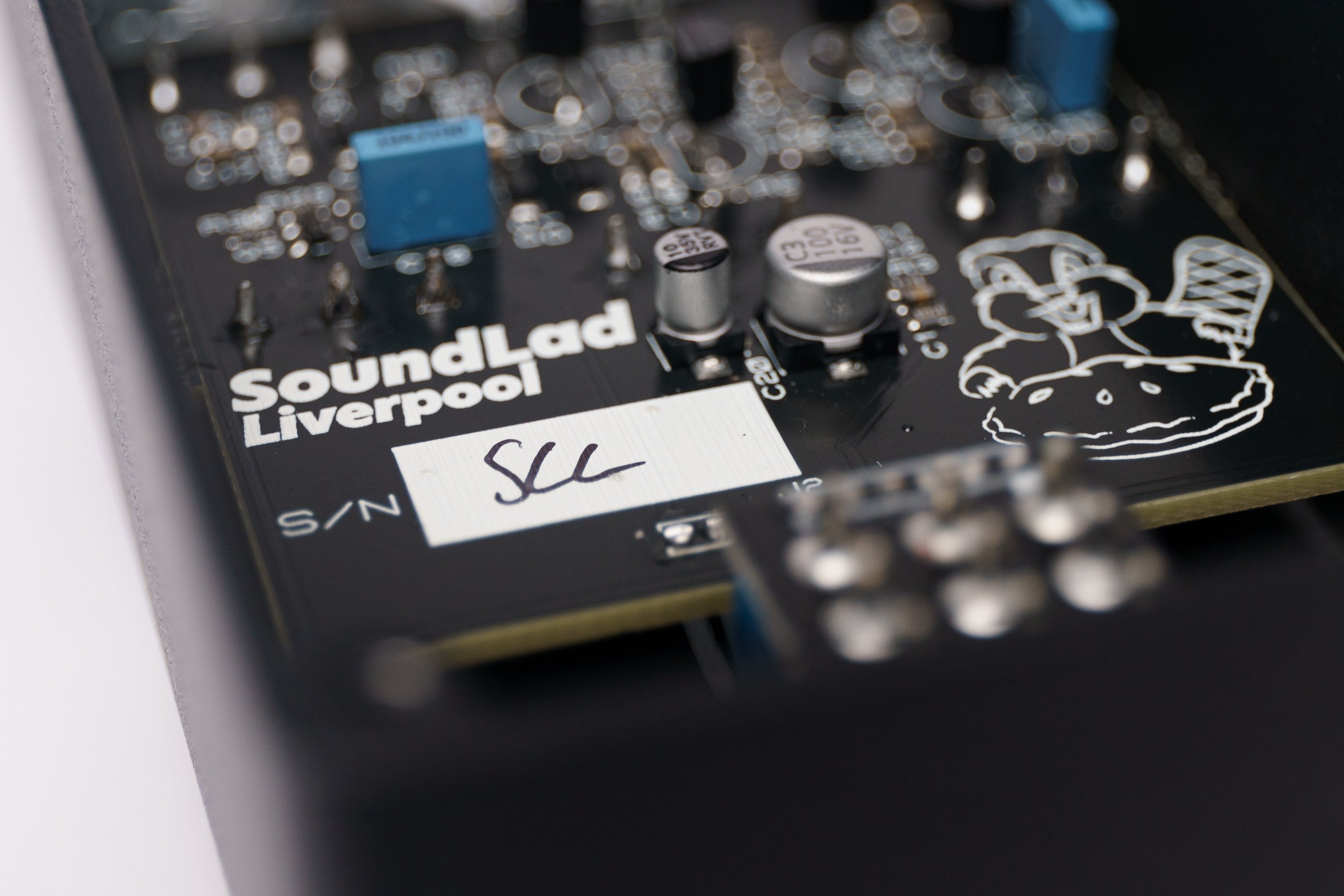 Close-up of an electronic circuit board with the words 'SoundLad Liverpool' printed on it, featuring capacitors, resistors, and a sticker with handwritten initials 'SLC', and a drawn image of a mouse with a pizza.