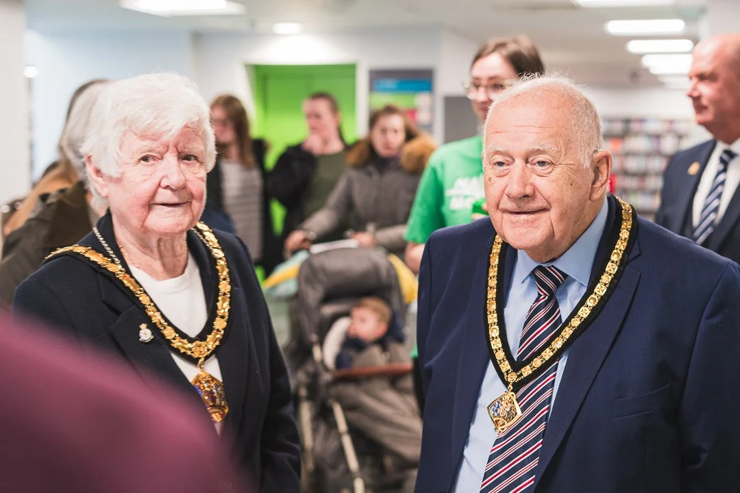 Two elderly individuals wearing ceremonial chain necklaces at a public event, surrounded by several people in a library or community center.