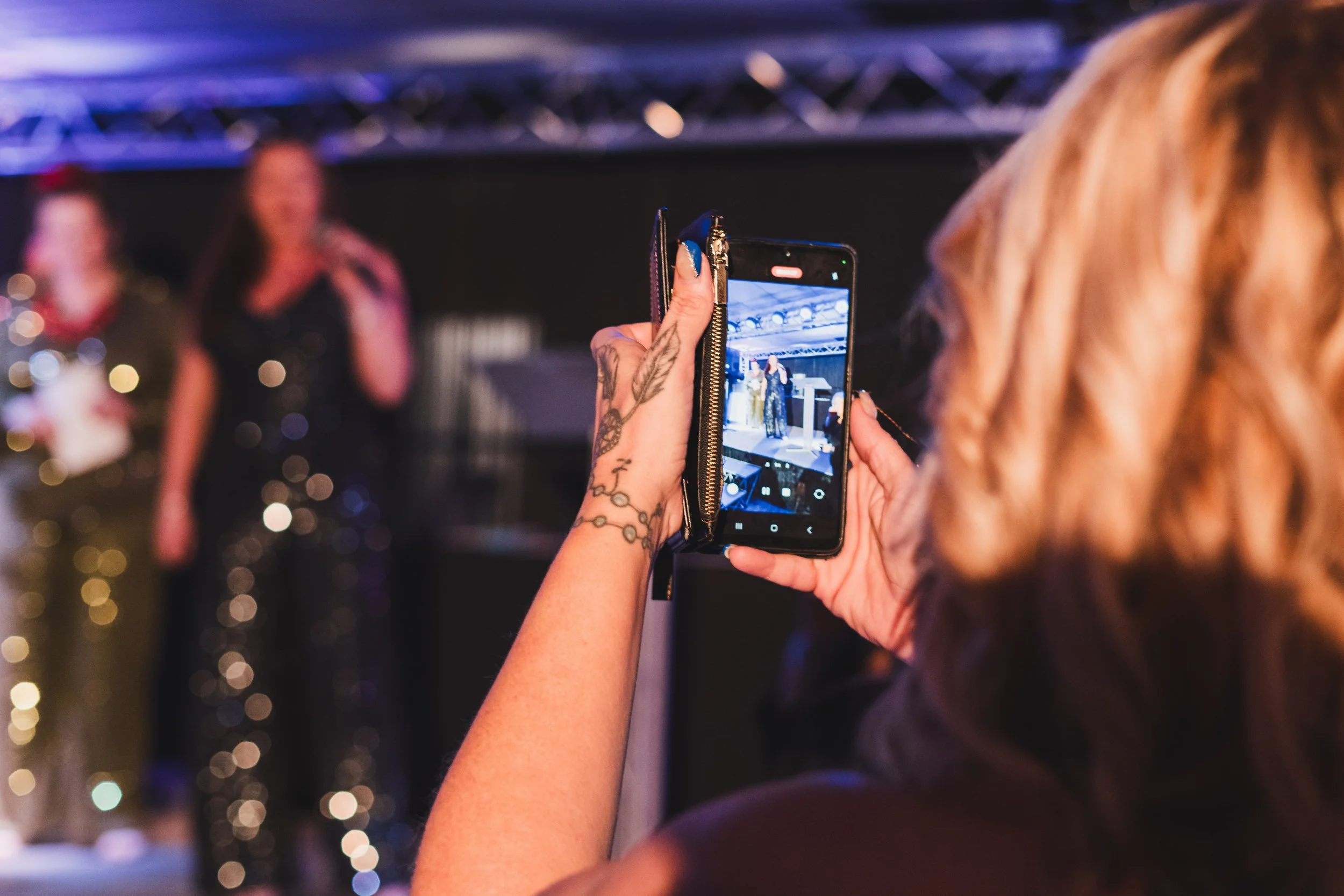 A woman with tattoos on her arm is taking a photo of a stage with two women standing on it using her smartphone at an indoor event.