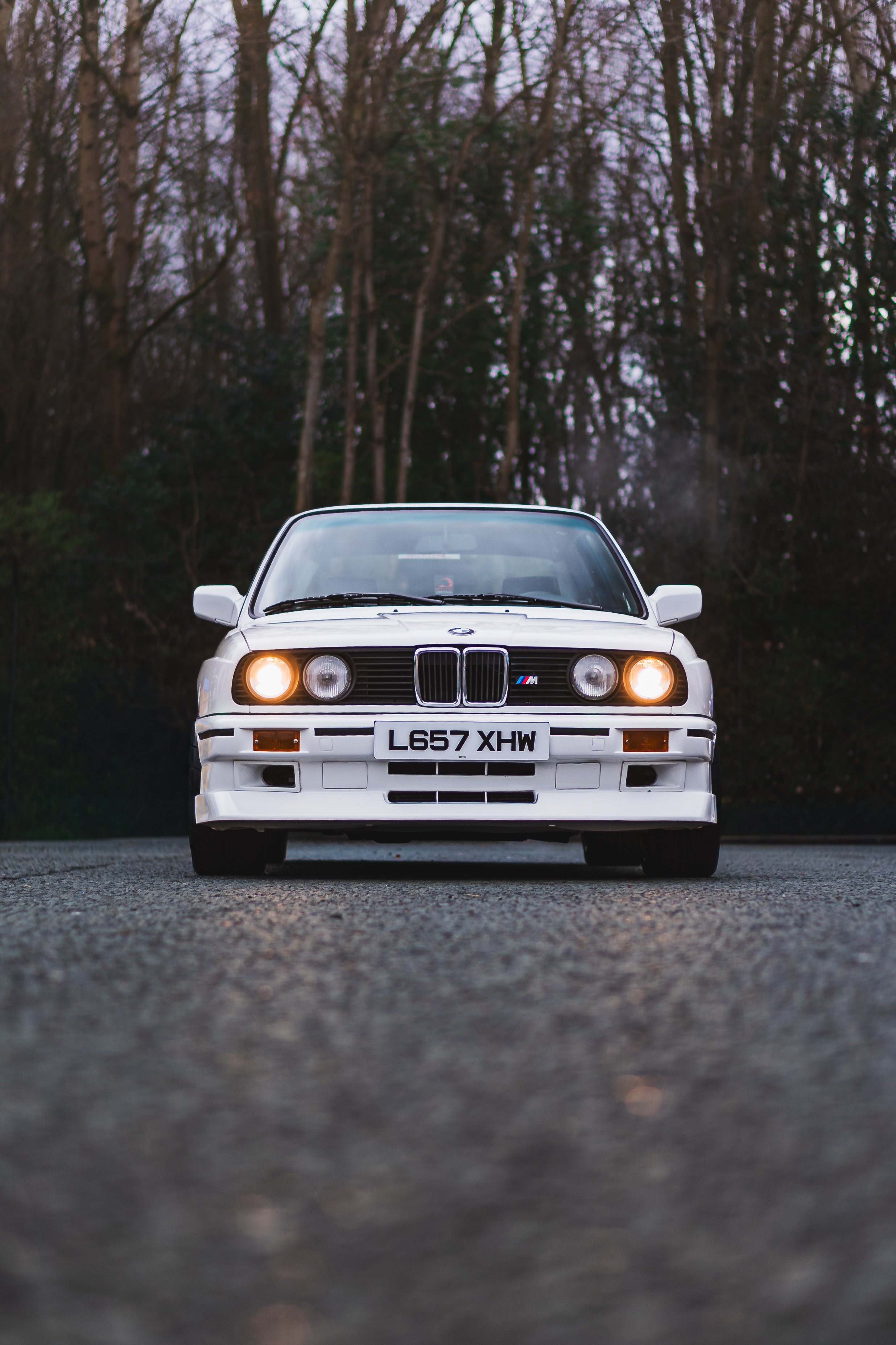 Front view of a white vintage BMW E30 M3 car on a gravel road with trees in the background.