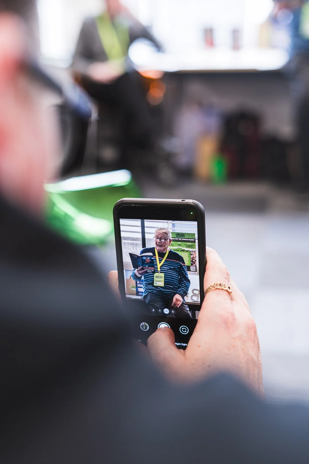 Person holding a smartphone shows a woman with glasses and a striped sweater on the screen, reading a booklet at an event, with a yellow lanyard and badge around her neck.