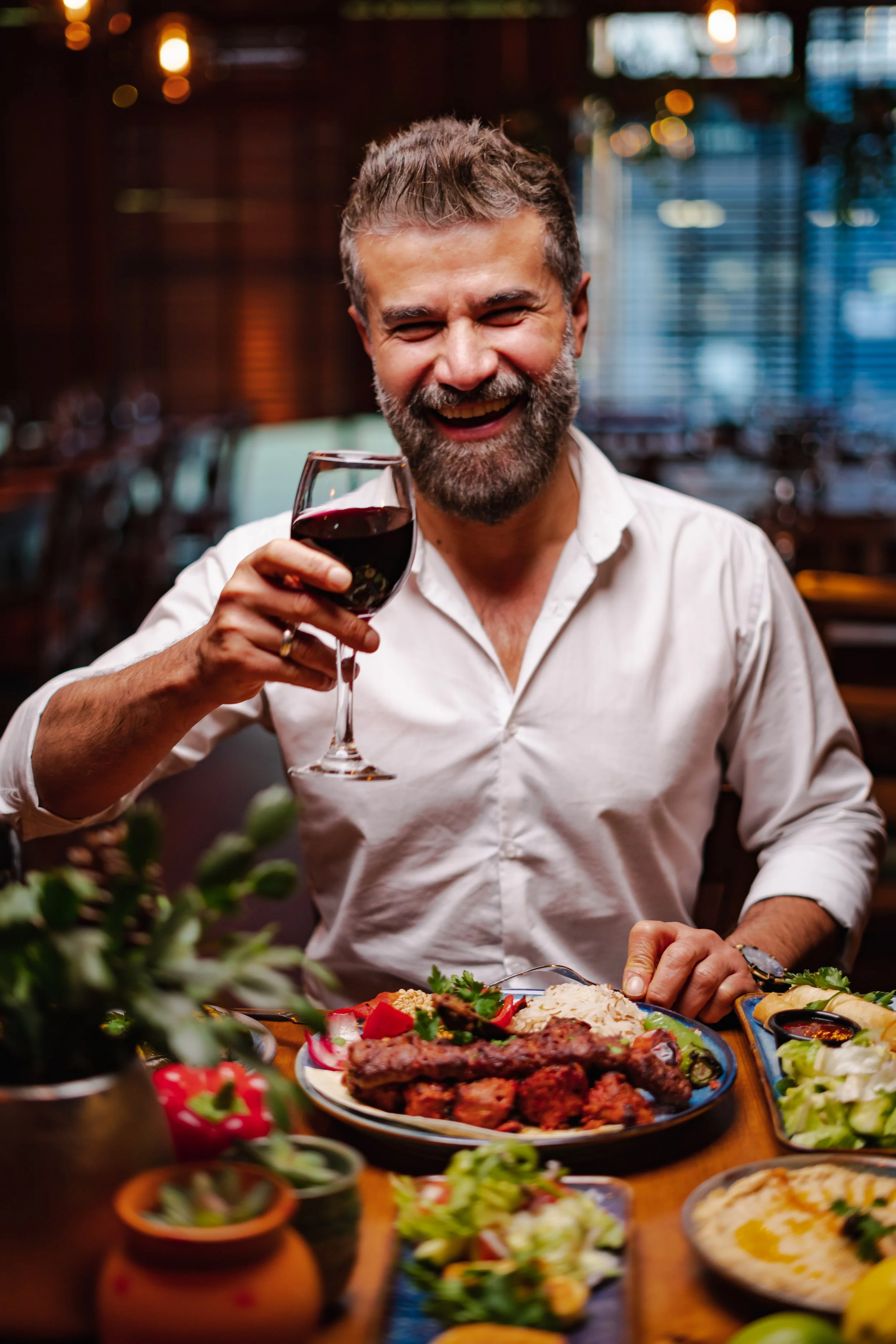A man with a beard laughing and holding a glass of red wine at a dinner table with various foods, including a plate of grilled meat, rice, salad, and other dishes, in a cozy restaurant.