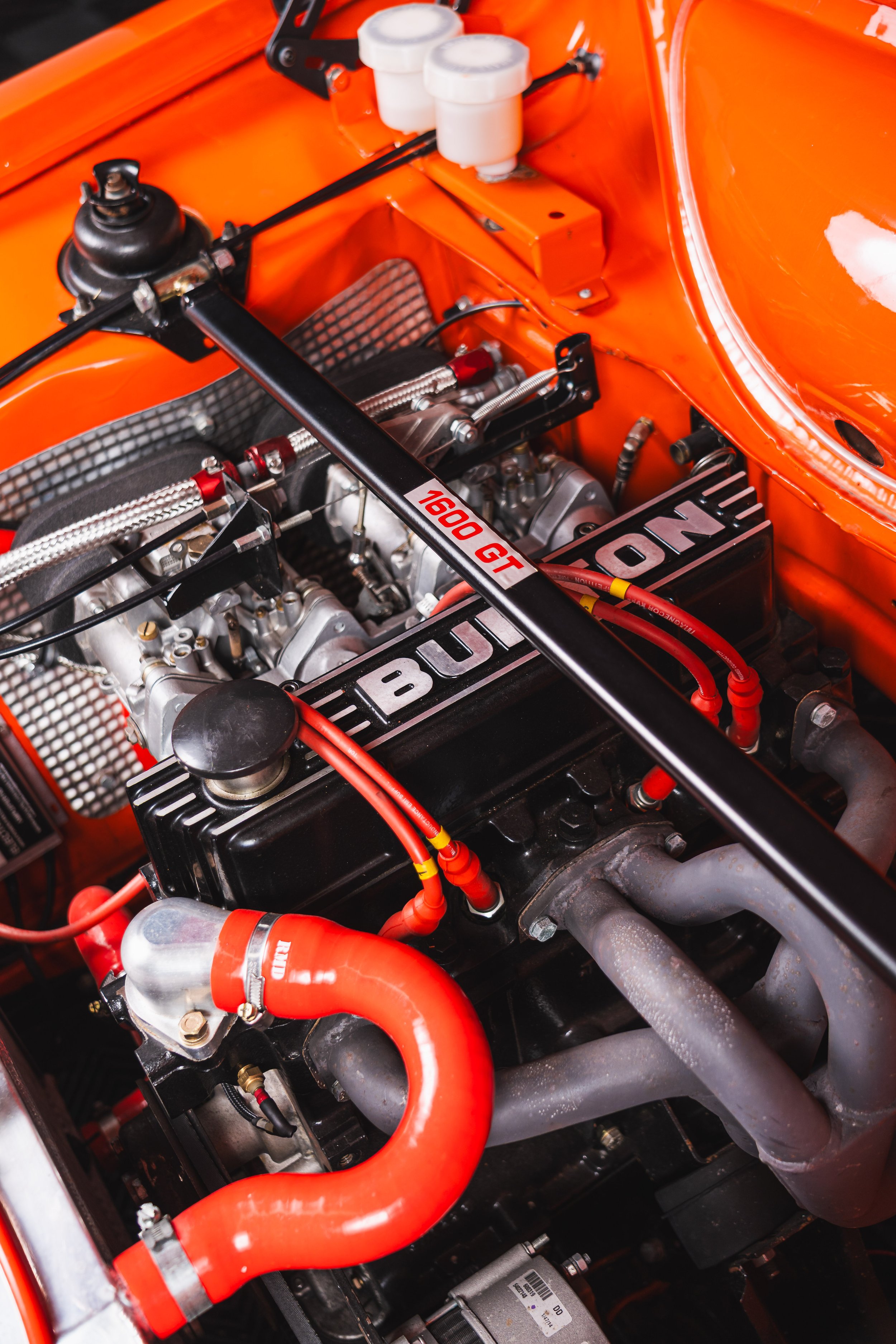Close-up view of a race car engine bay, showcasing a high-performance engine labeled 'BUN.' Red hoses and wires are connected to various engine components, with an orange-painted engine bay and a black GT Spec piping.