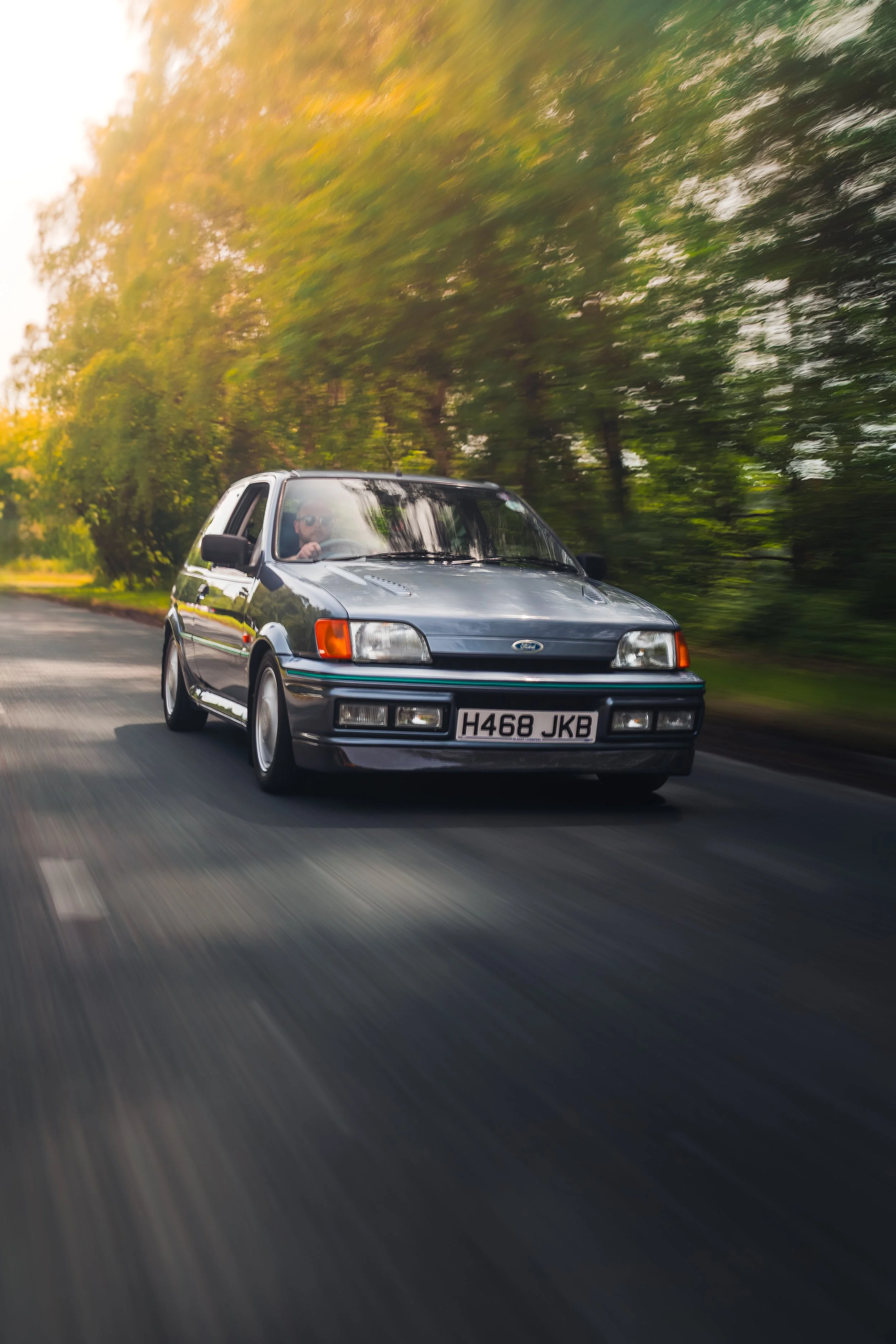 A silver classic hatchback car with a visible license plate 'H468 JKB' driving on a road surrounded by greenery and trees, with motion blur indicating speed.