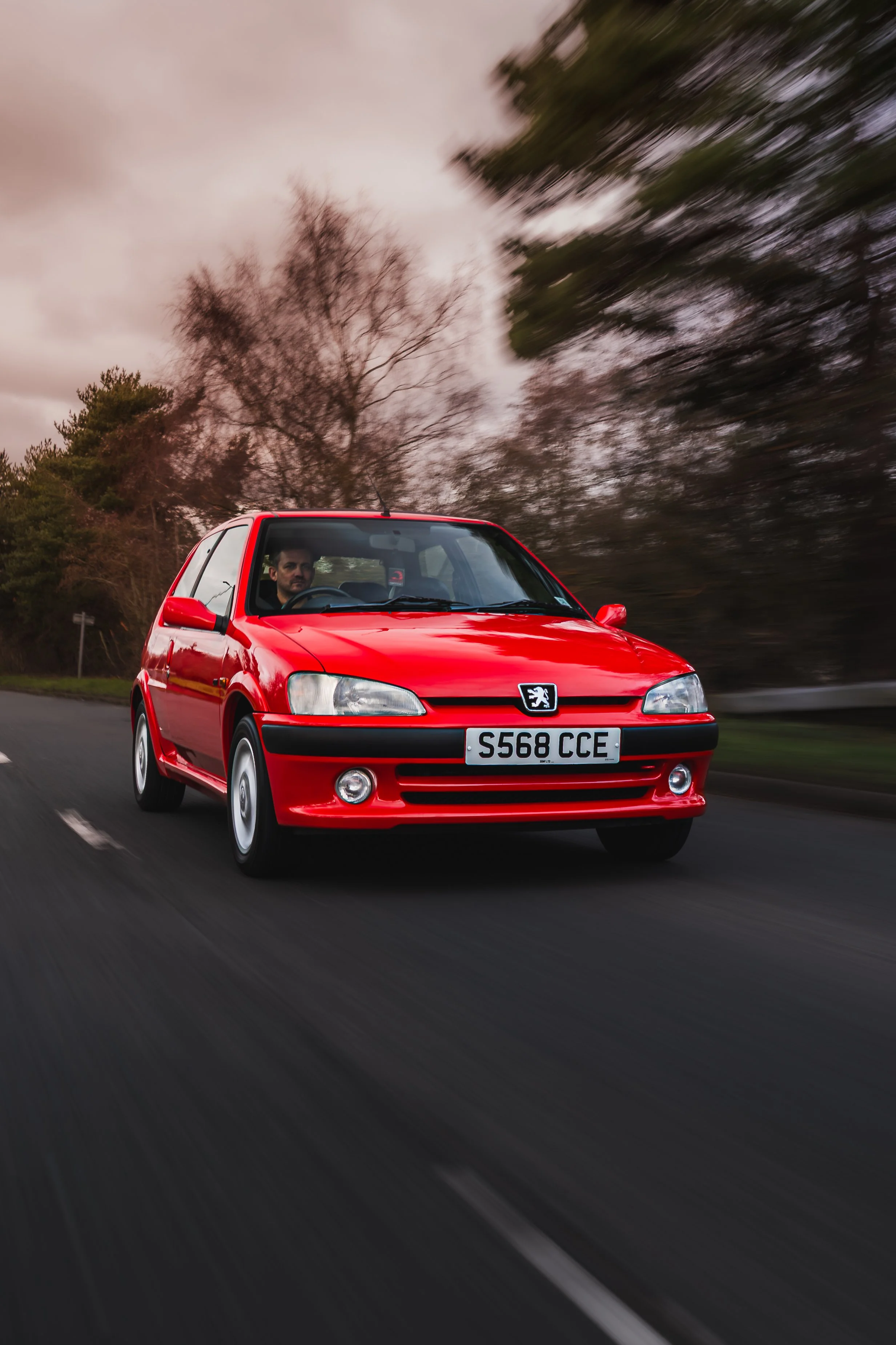 A red Peugeot hatchback driving on a road with a driver visible through the windshield, with blurred trees and cloudy sky in the background.
