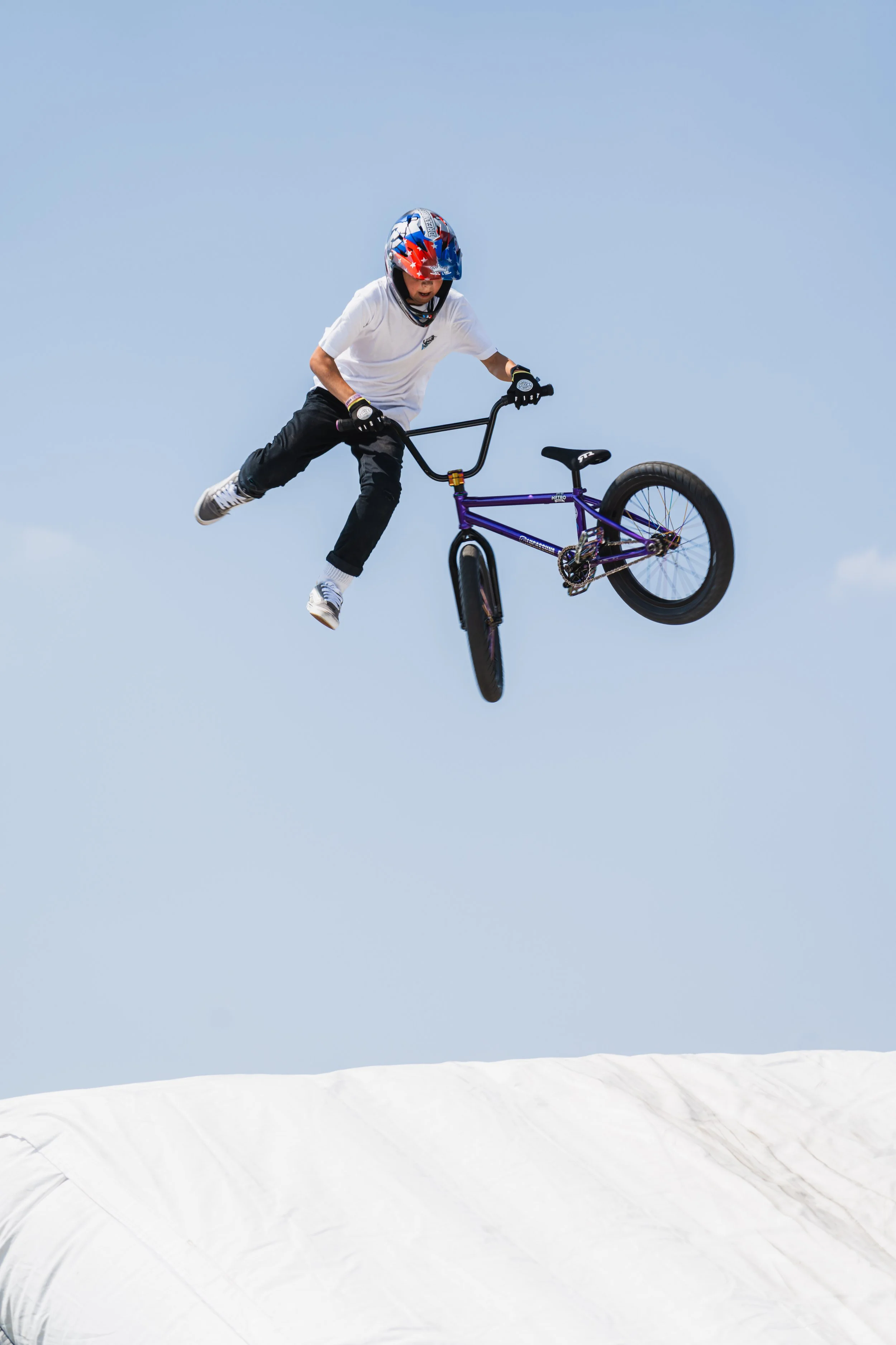 A person performing a BMX stunt in mid-air against a blue sky, dressed in casual clothes and wearing a helmet, with a white inflatable ramp below.