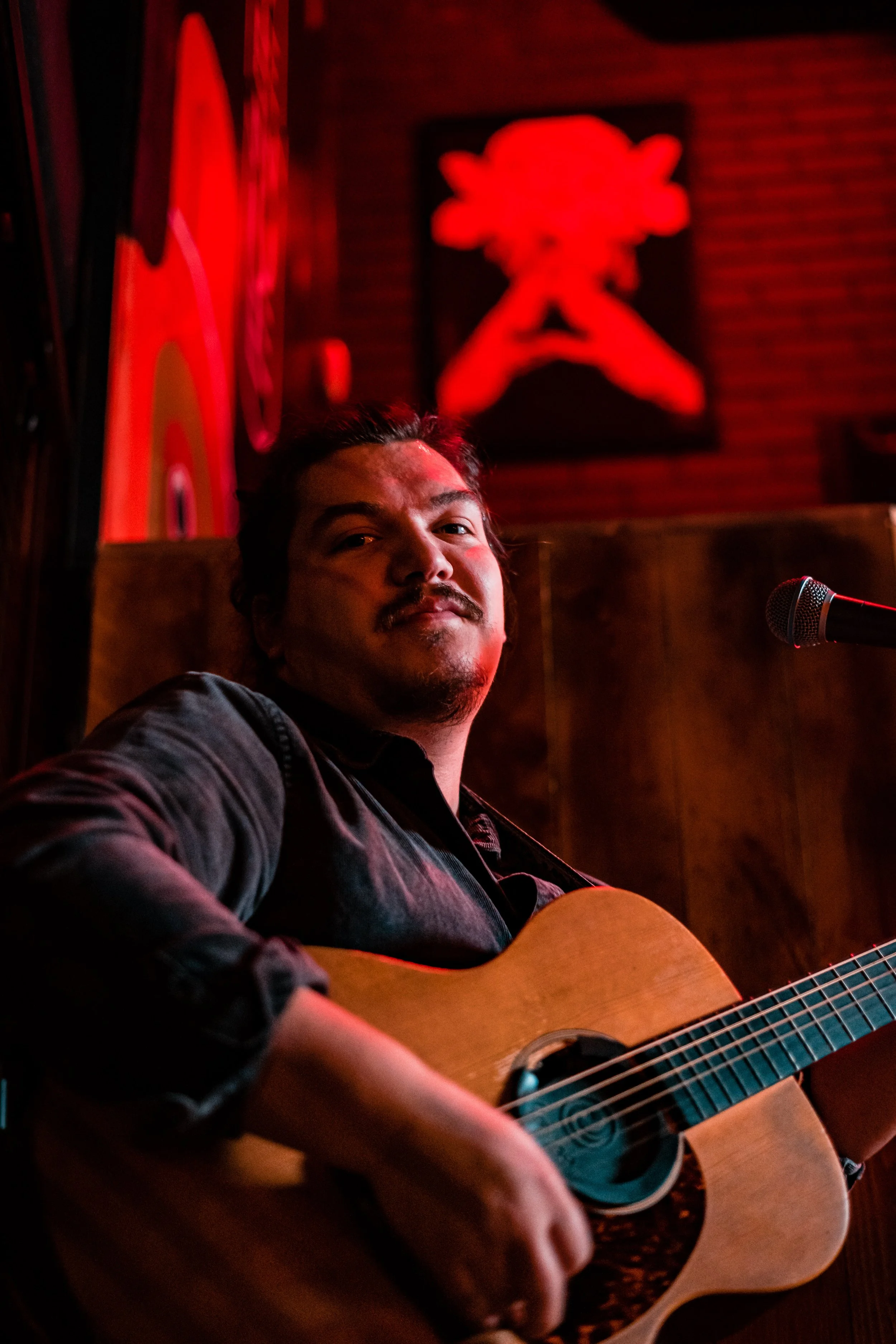 A man playing an acoustic guitar on stage in a dimly lit venue, with red neon lighting and abstract artwork in the background.