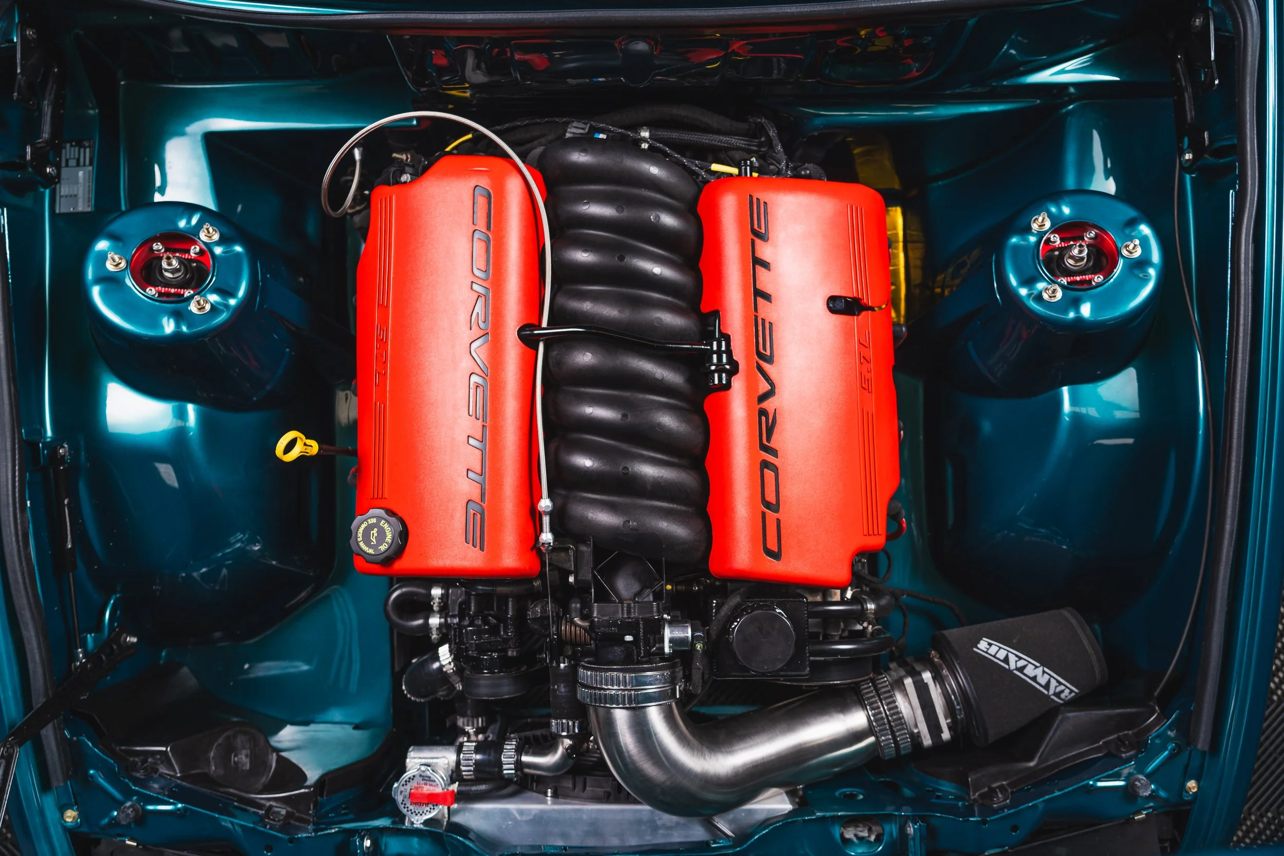 Close-up view of a car engine with red valve covers labeled 'Corvette' and 'LT2', black intake manifold, and a polished metal pipe, all inside a teal-colored engine bay.