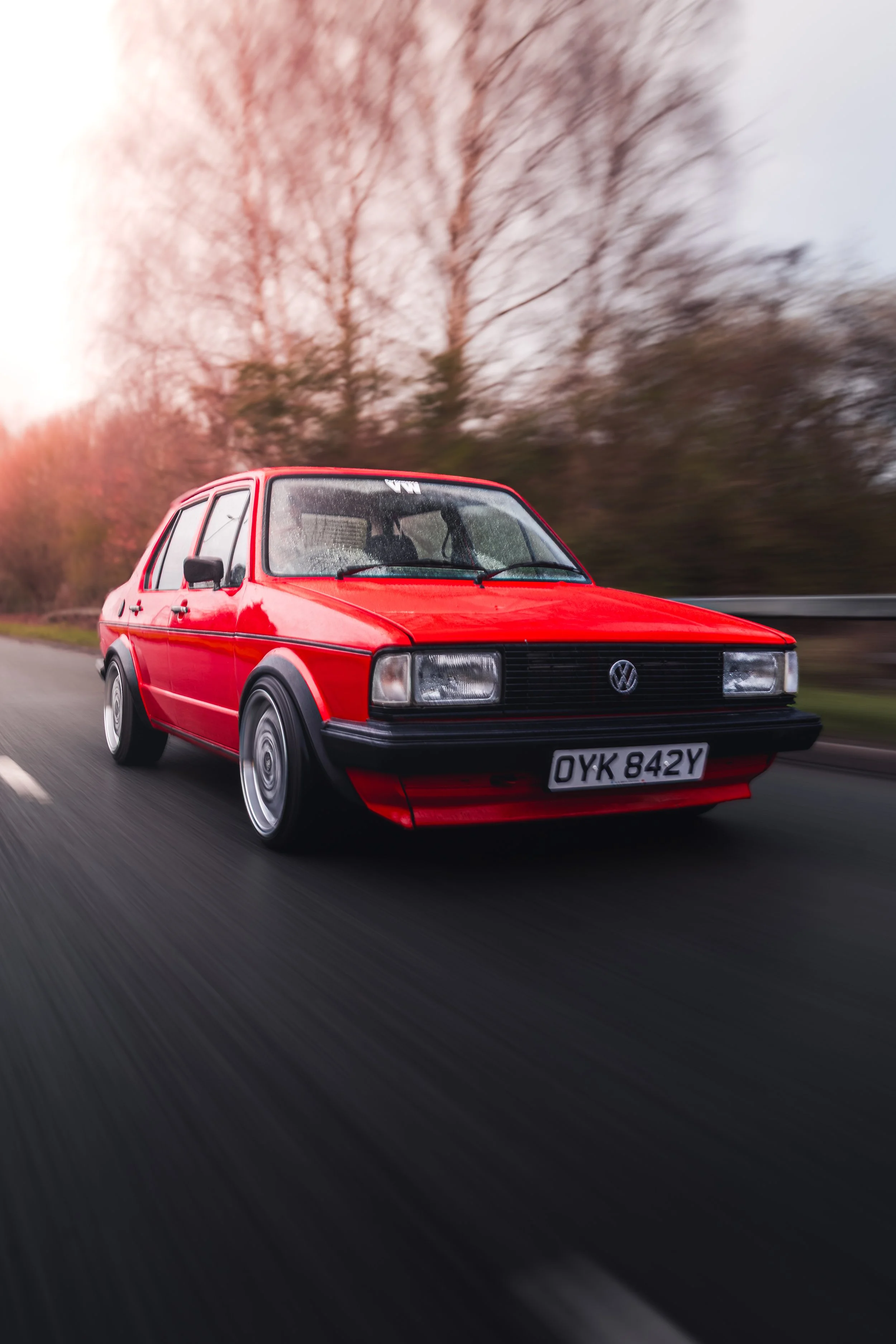 A red vintage Volkswagen hatchback car driving on a road during sunset with blurred trees in the background.