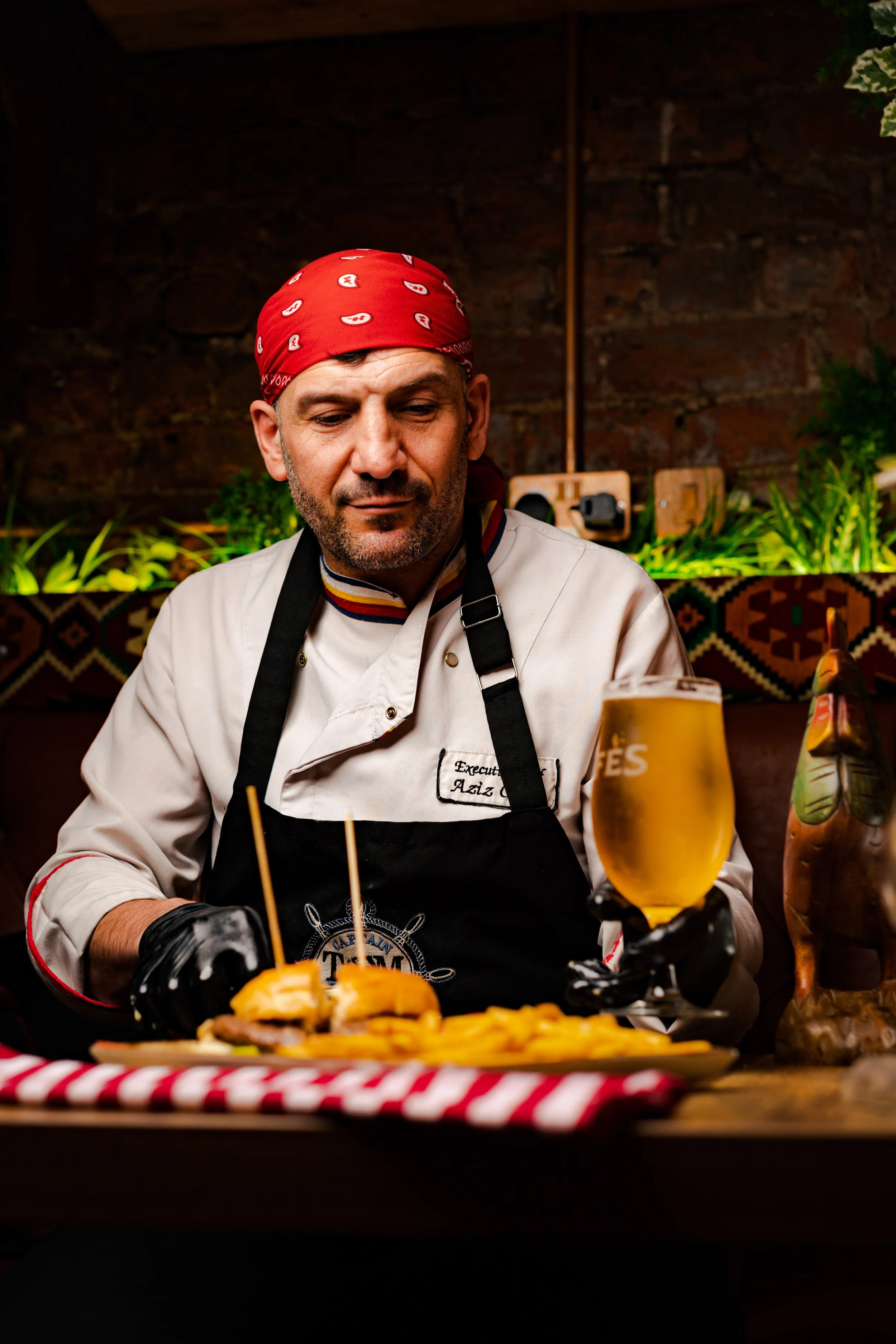 A chef dressed in a white chef's coat, red bandana, and black apron, sitting at a table with a plate of sliders and fries, holding a glass of beer in a restaurant setting.