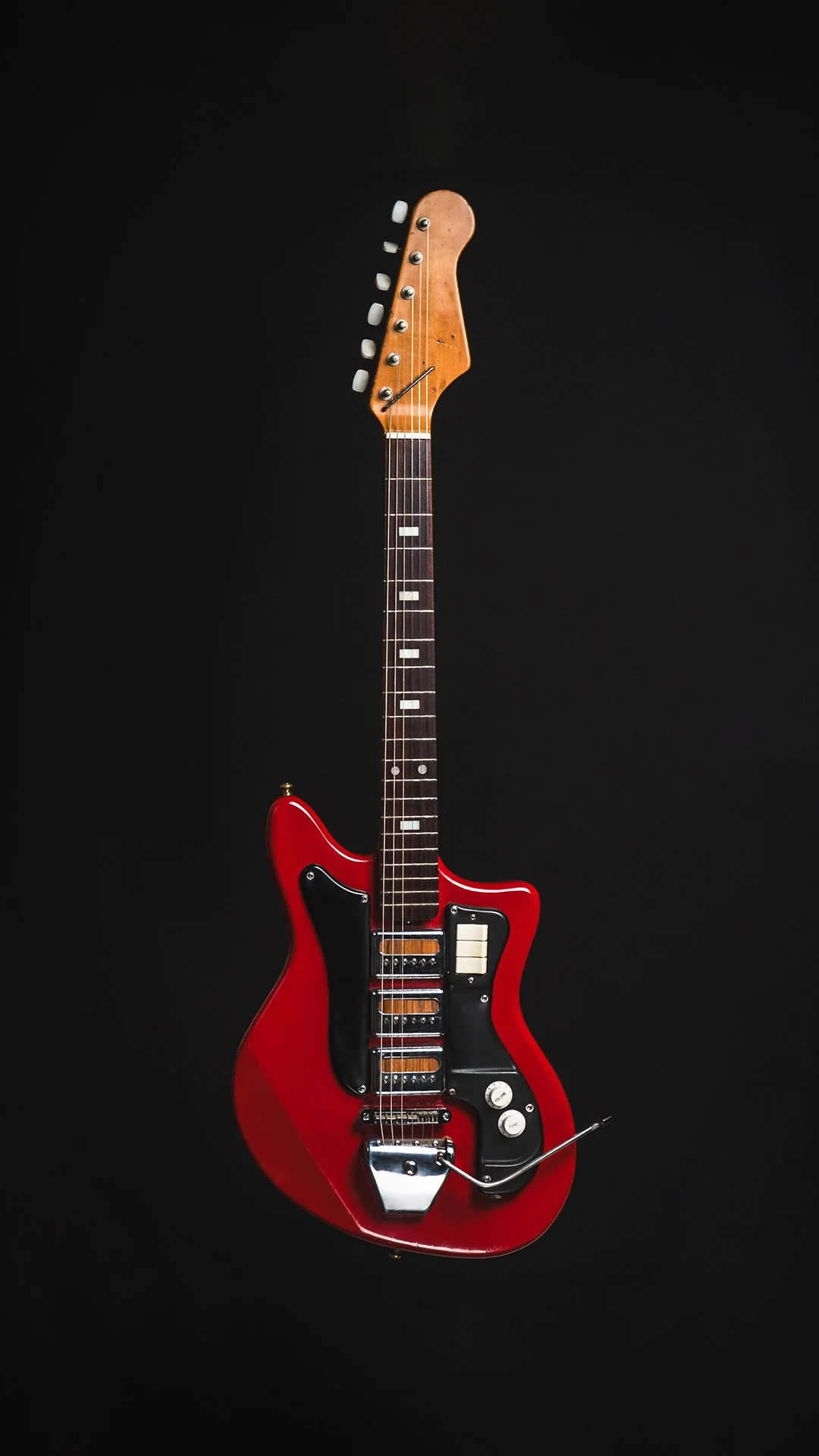 A vintage red and black electric guitar with a unique body shape, featuring a single coil pickup, a black pickguard, and a whammy bar, against a black background.