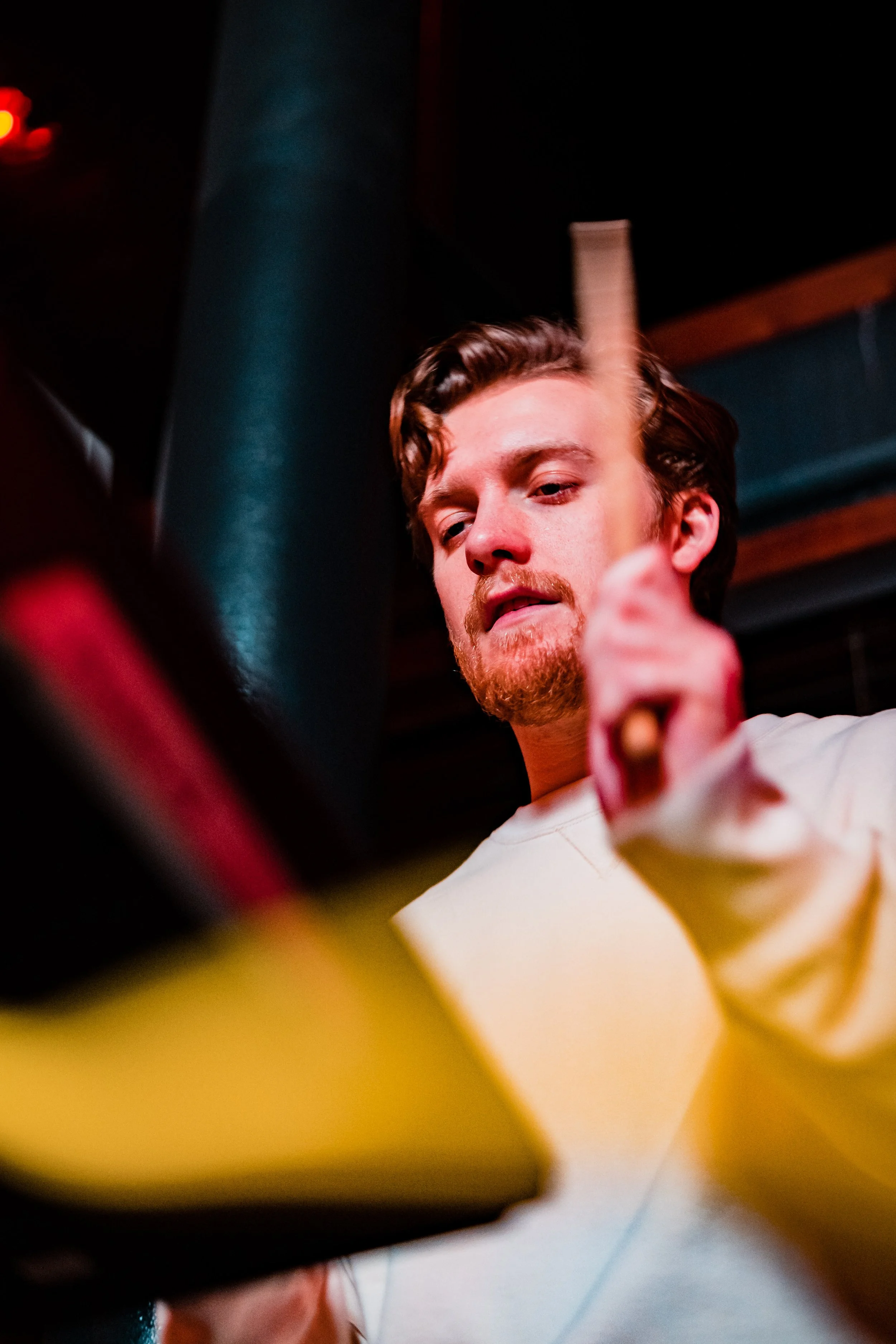 Young man with brown hair and beard pointing at a book or tablet with colorful covers on a table, indoors.