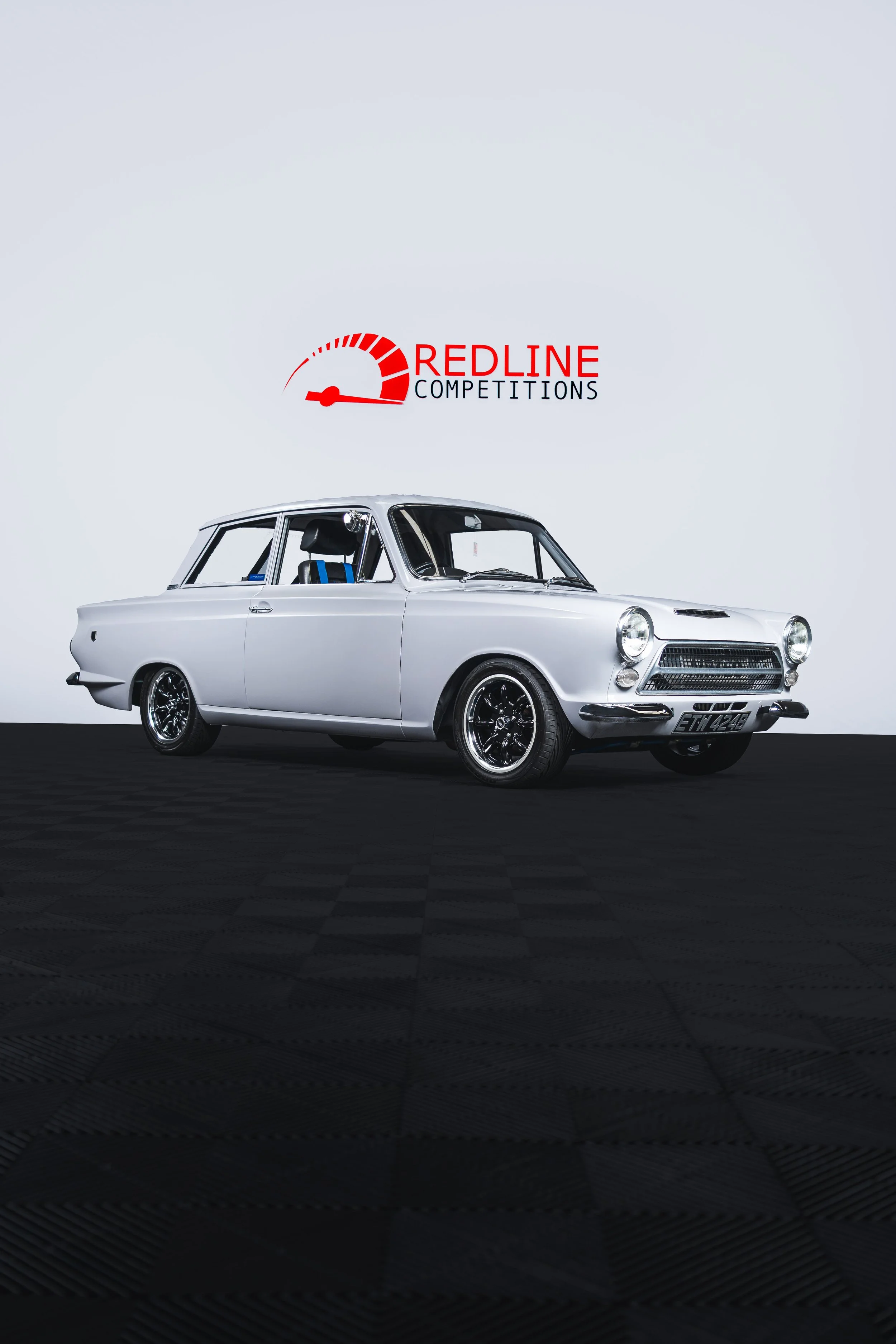 A classic silver car on display at a competition with a 'Redline Competitions' sign in the background.