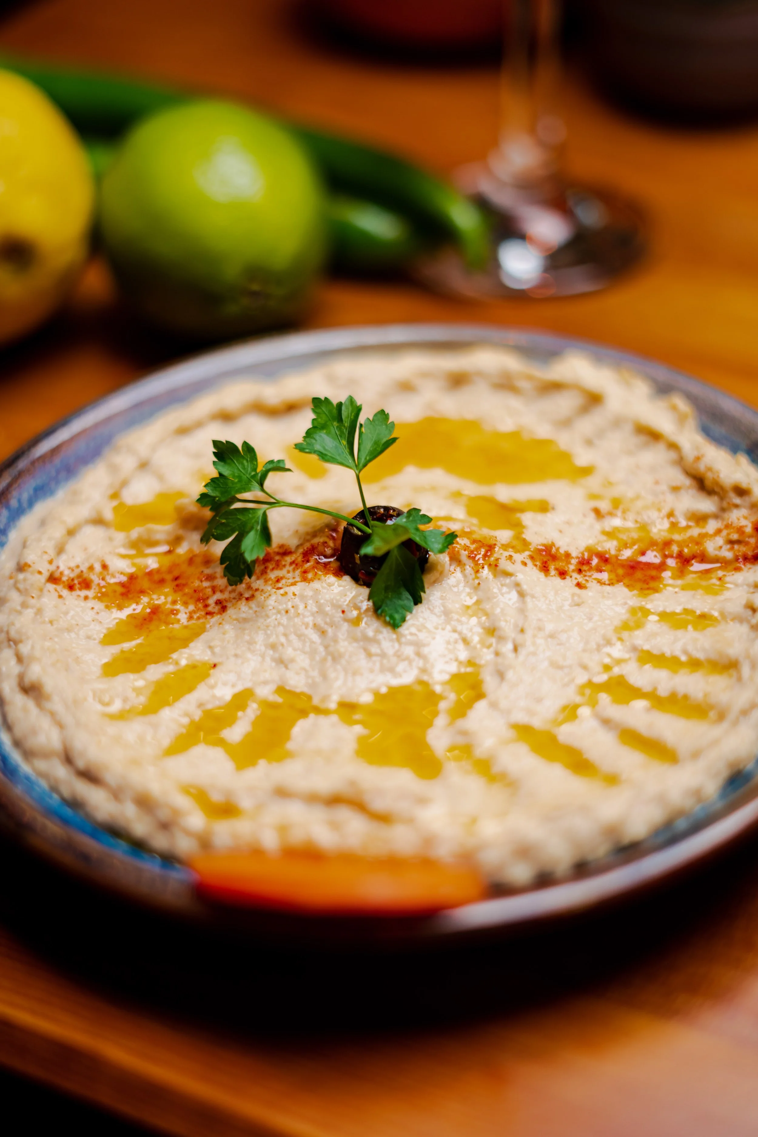 A bowl of hummus garnished with parsley, olive, and drizzle of olive oil, with lemons, avocado, jalapenos, wine glass in the background on a wooden table.