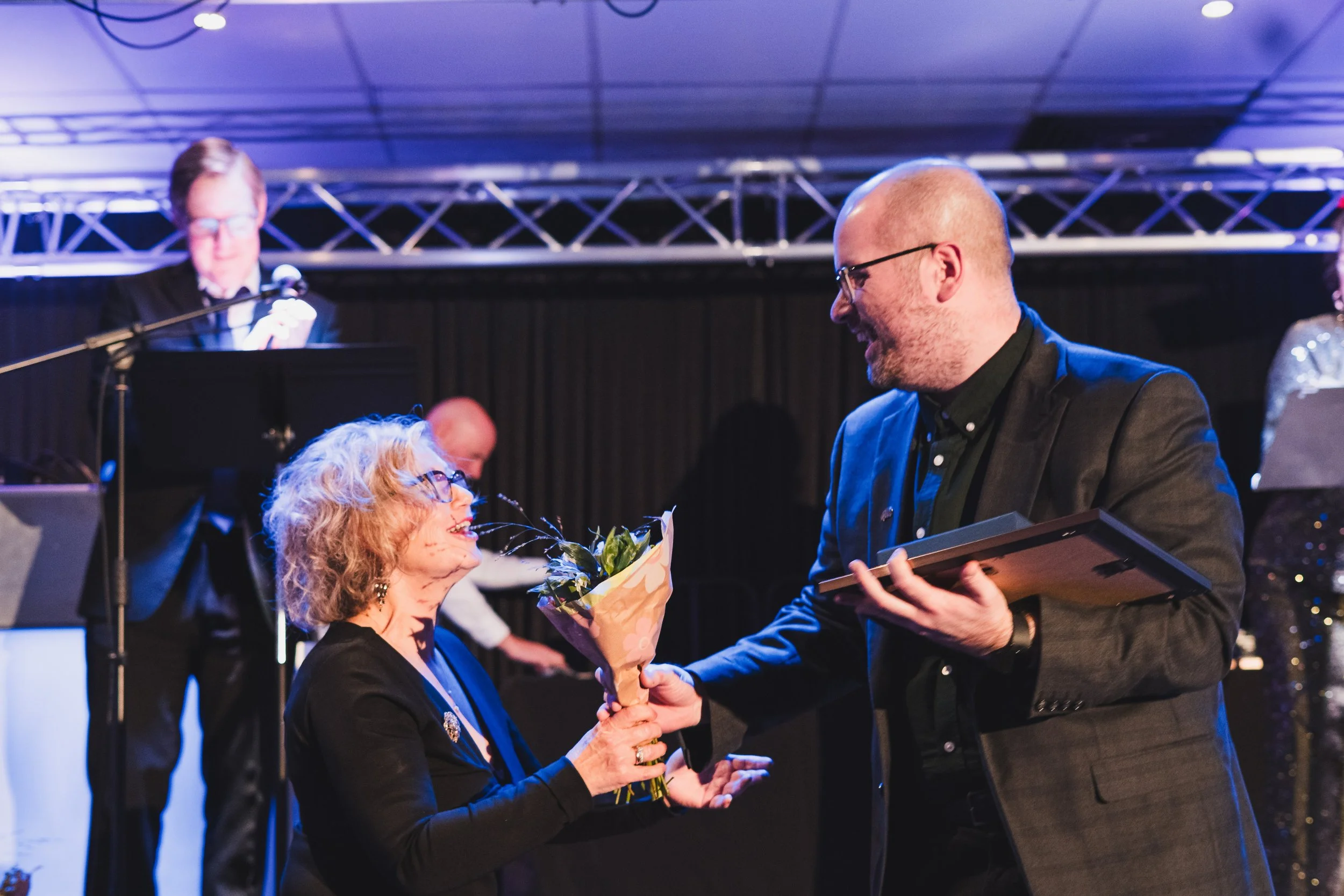 A woman with curly gray hair and glasses receives a bouquet of flowers from a man with glasses and a beard, both smiling, at an indoor event with a stage, musical equipment, and a singer in the background.