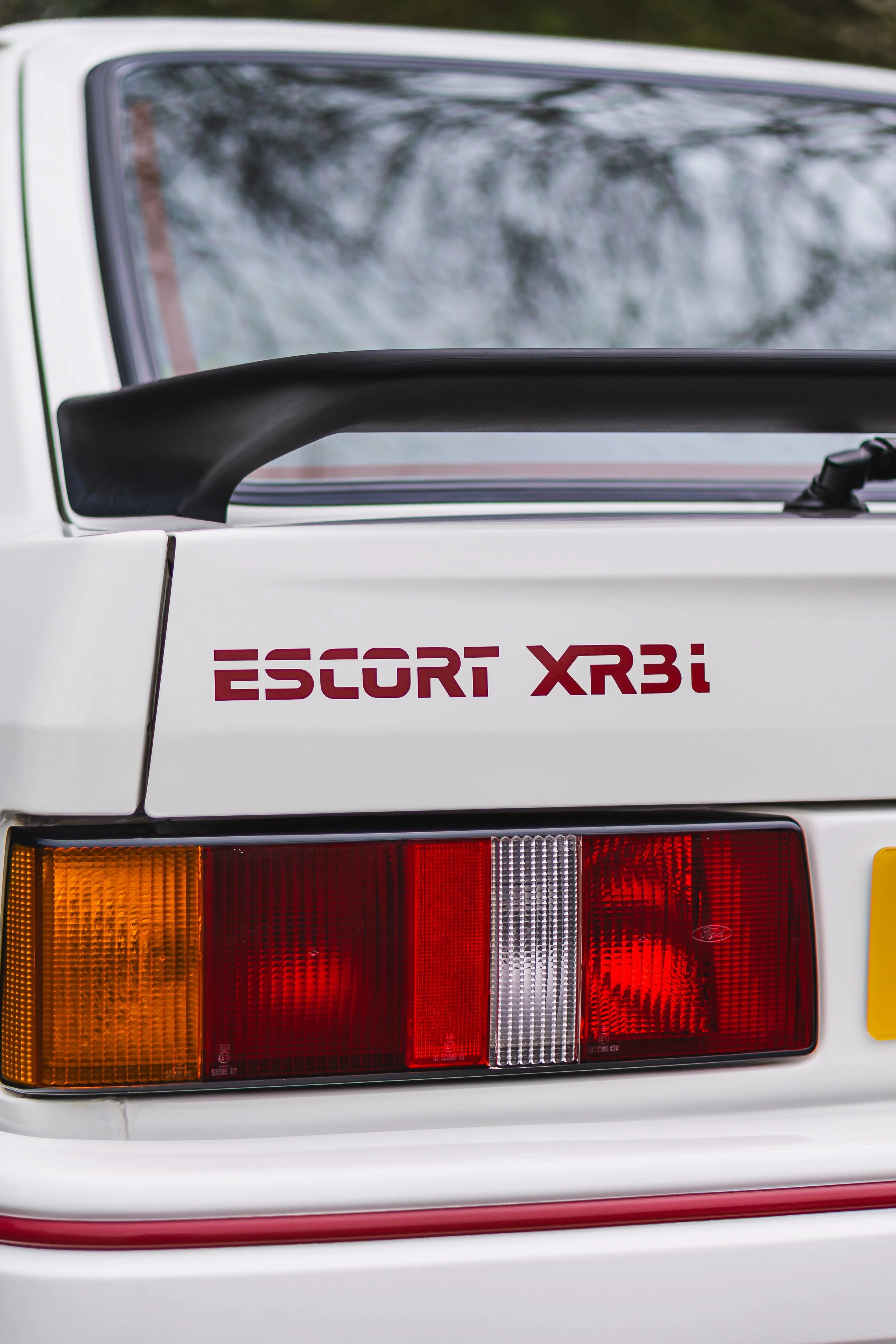 Close-up of the rear end of a white Ford Escort XR3i, showing the tail lights, rear window, and a decal that reads 'ESCORT XR3i'.