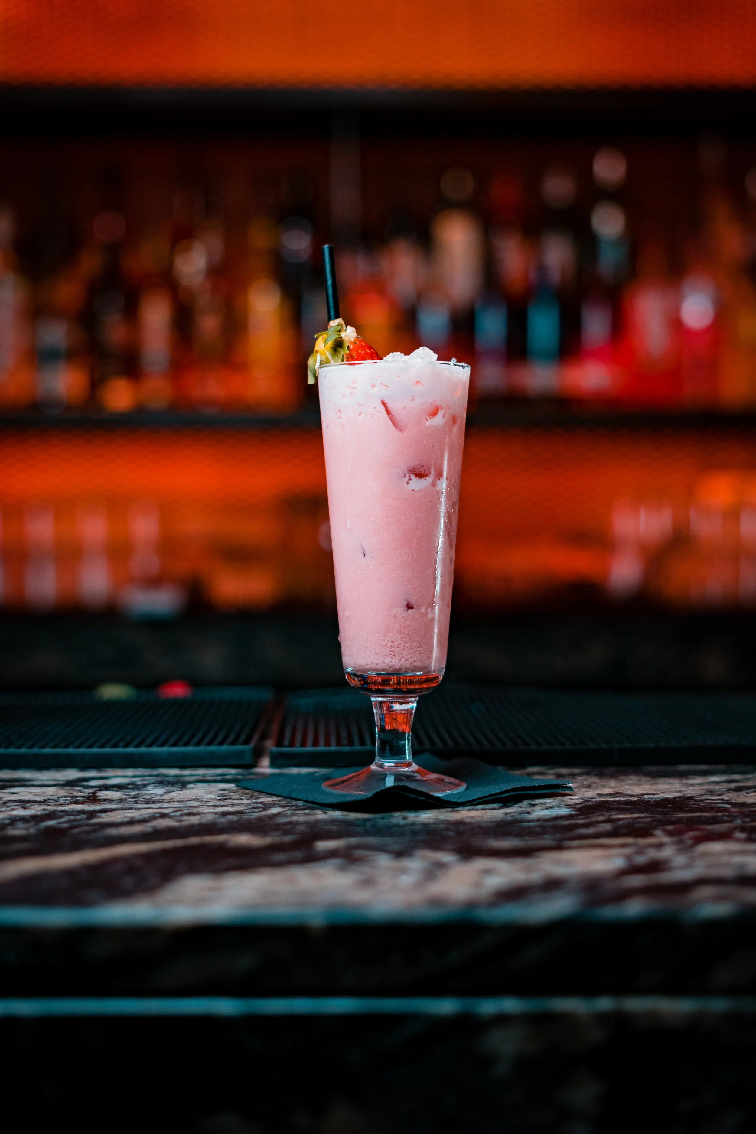 Pink strawberry cocktail in a tall glass with a straw, garnished with a strawberry and a fruit slice, on a bar counter with a blurred bar backdrop.