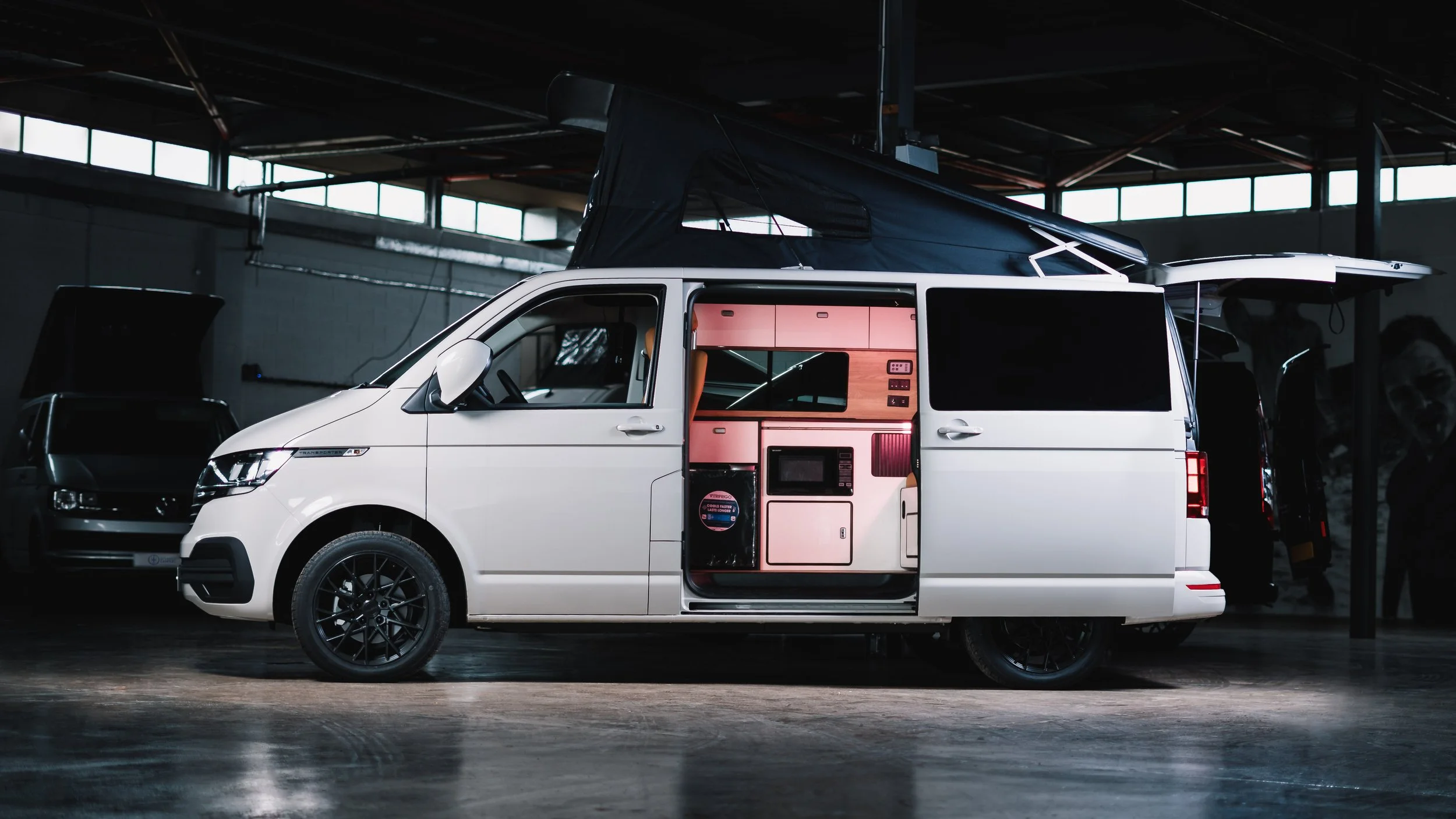 White camper van with a pop-up roof and custom interior, parked indoors at a vehicle show.