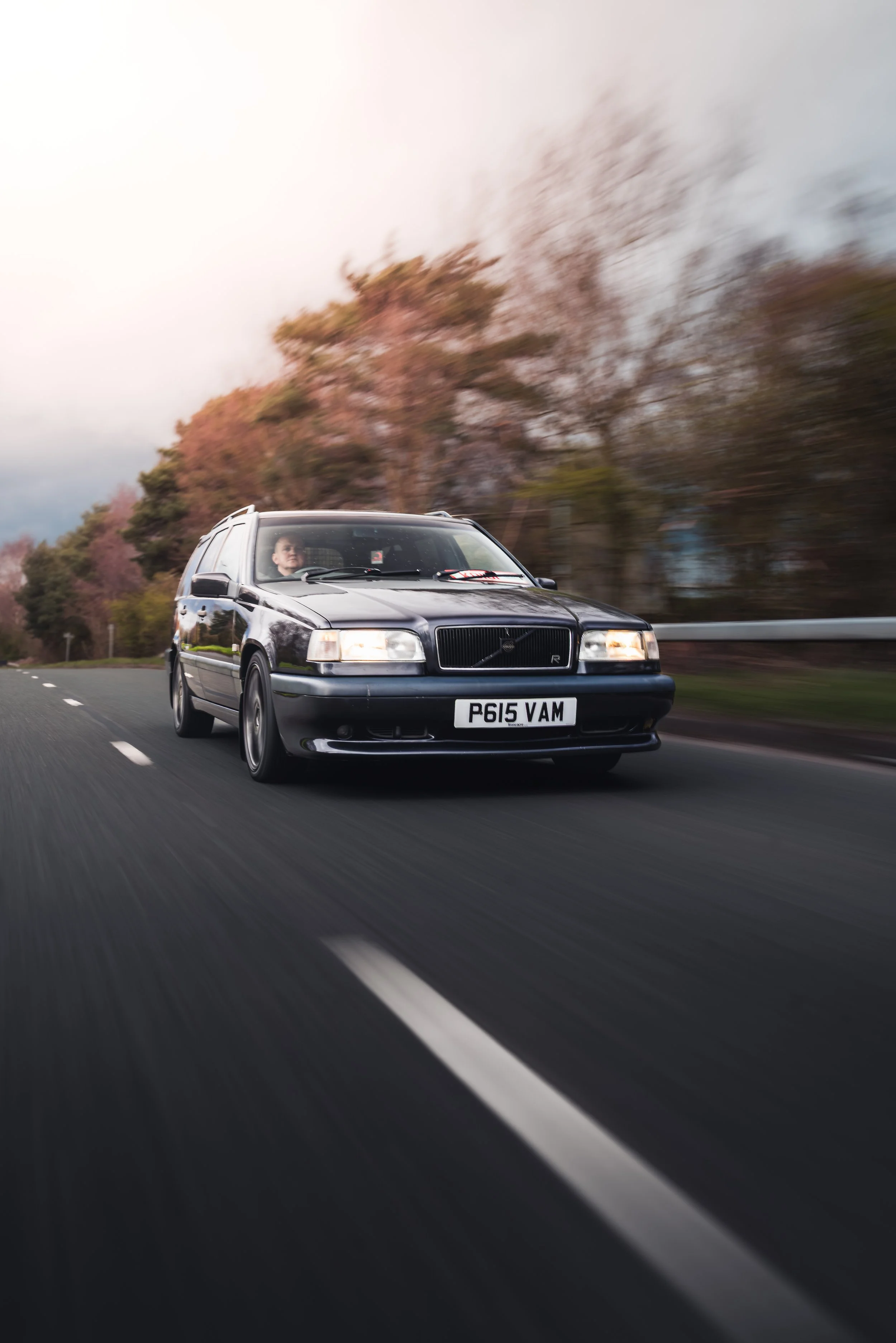 A black vintage Volvo station wagon with the license plate 'P615 VAM' driving on a highway with blurred trees and sky in the background, indicating motion.