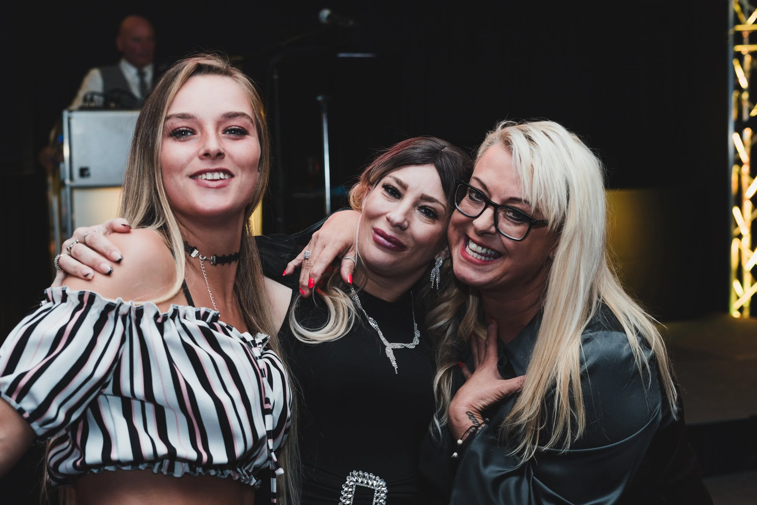 Three women smiling and hugging each other at a social event with a person in the background playing music.