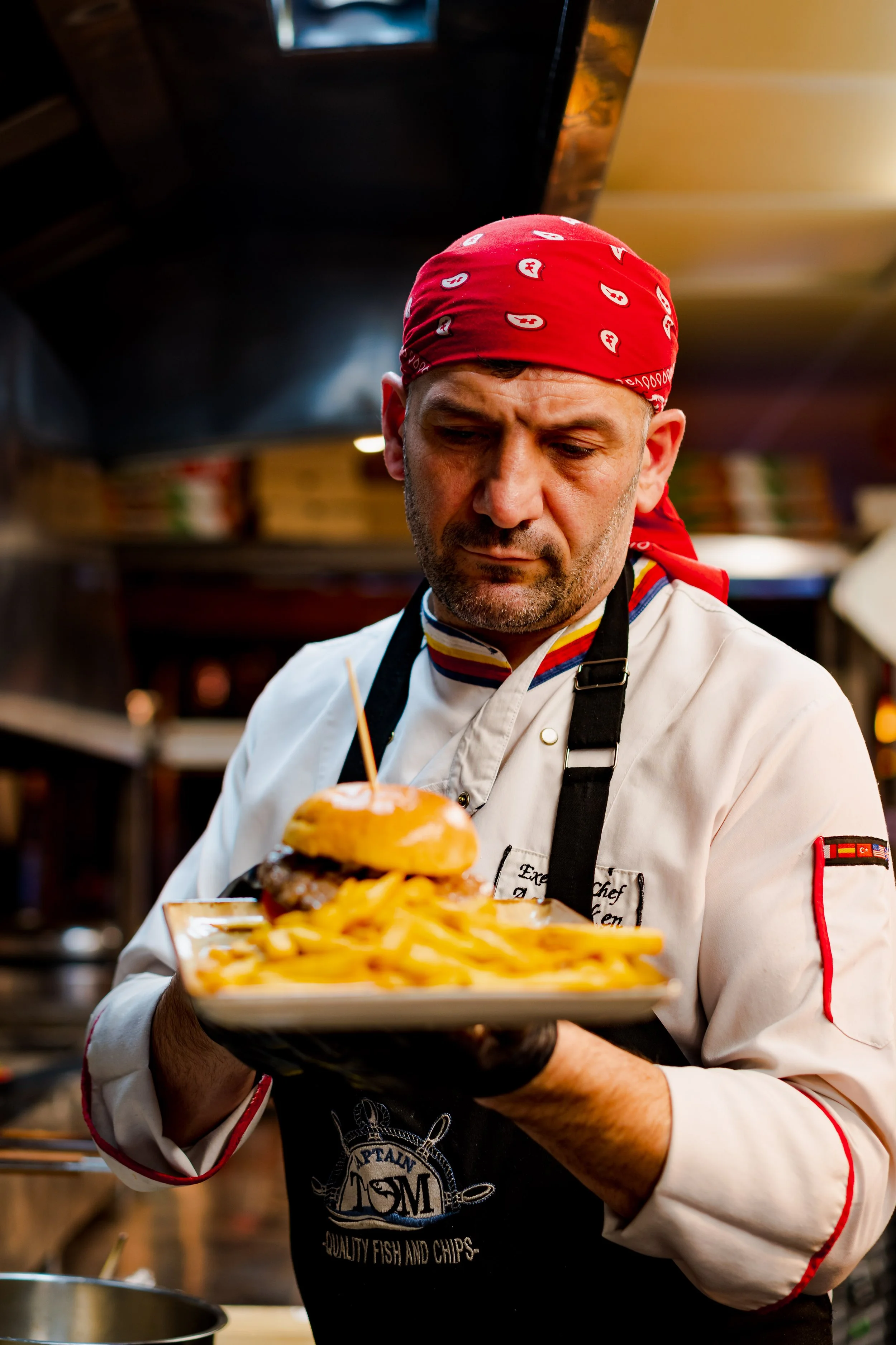 A chef in a red bandana holding a tray with a burger and French fries inside a restaurant kitchen.