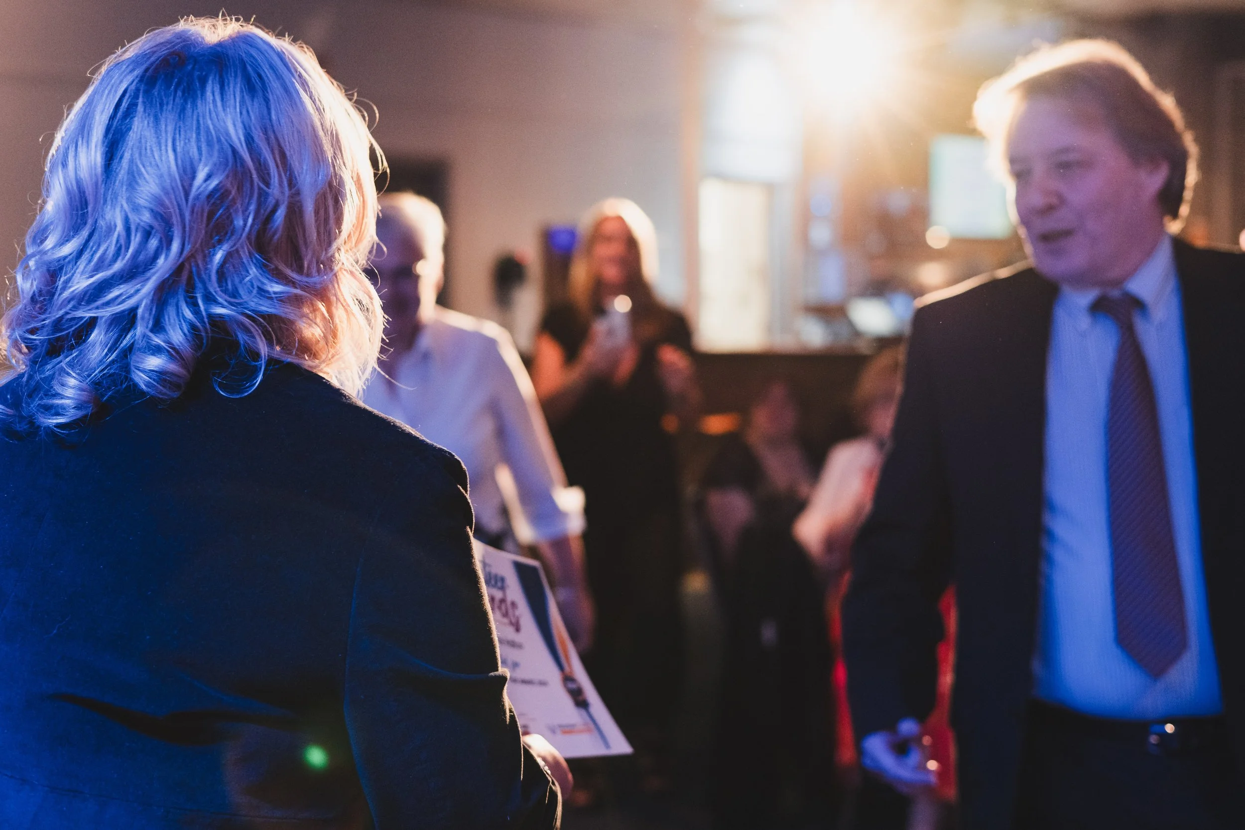 Two people, a woman with curly blonde hair and a man with blond hair, are in conversation at a social event. The background shows other attendees and warm lighting.