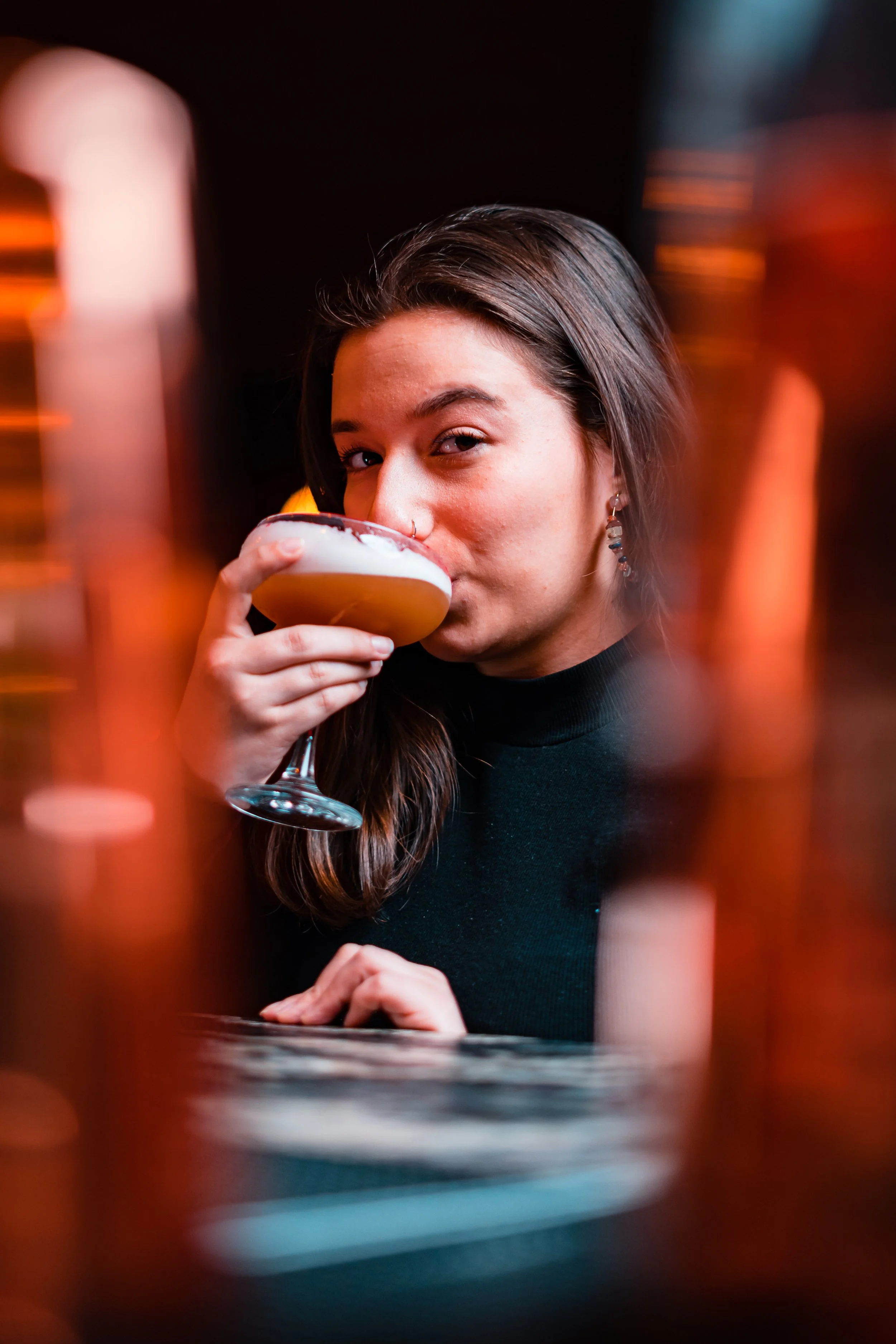 A woman with long brown hair, wearing a black top and earrings, is holding a cocktail glass filled with a drink topped with foam, and is taking a sip, with a blurred background.