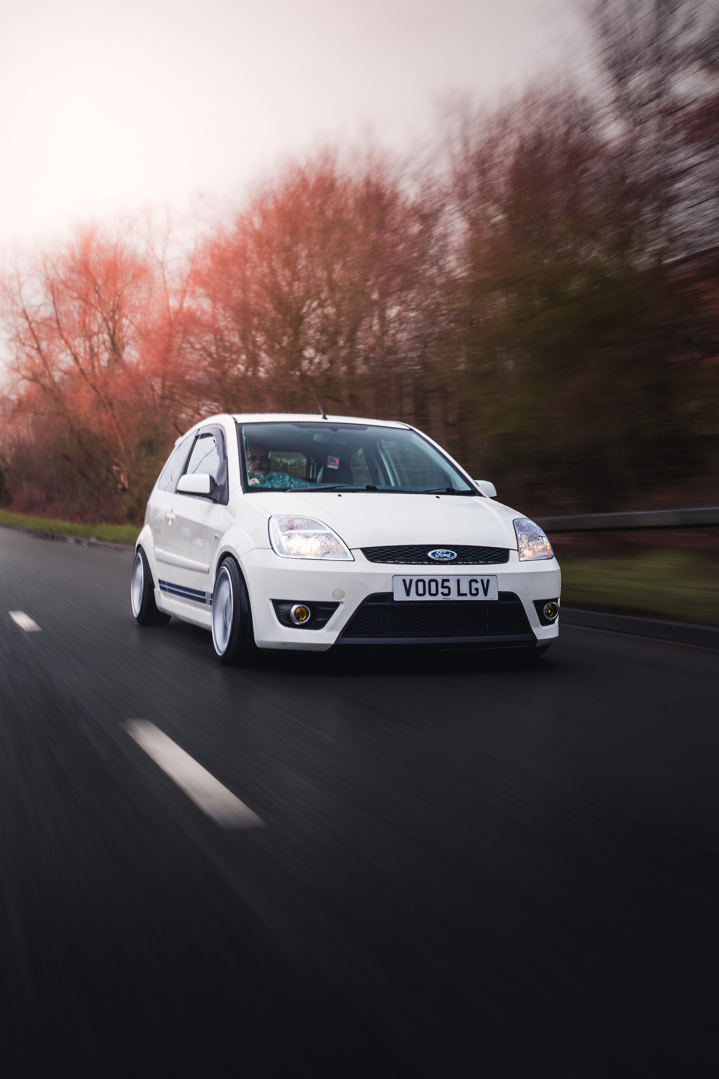 White Ford car driving on a highway with trees and a pinkish sky in the background.