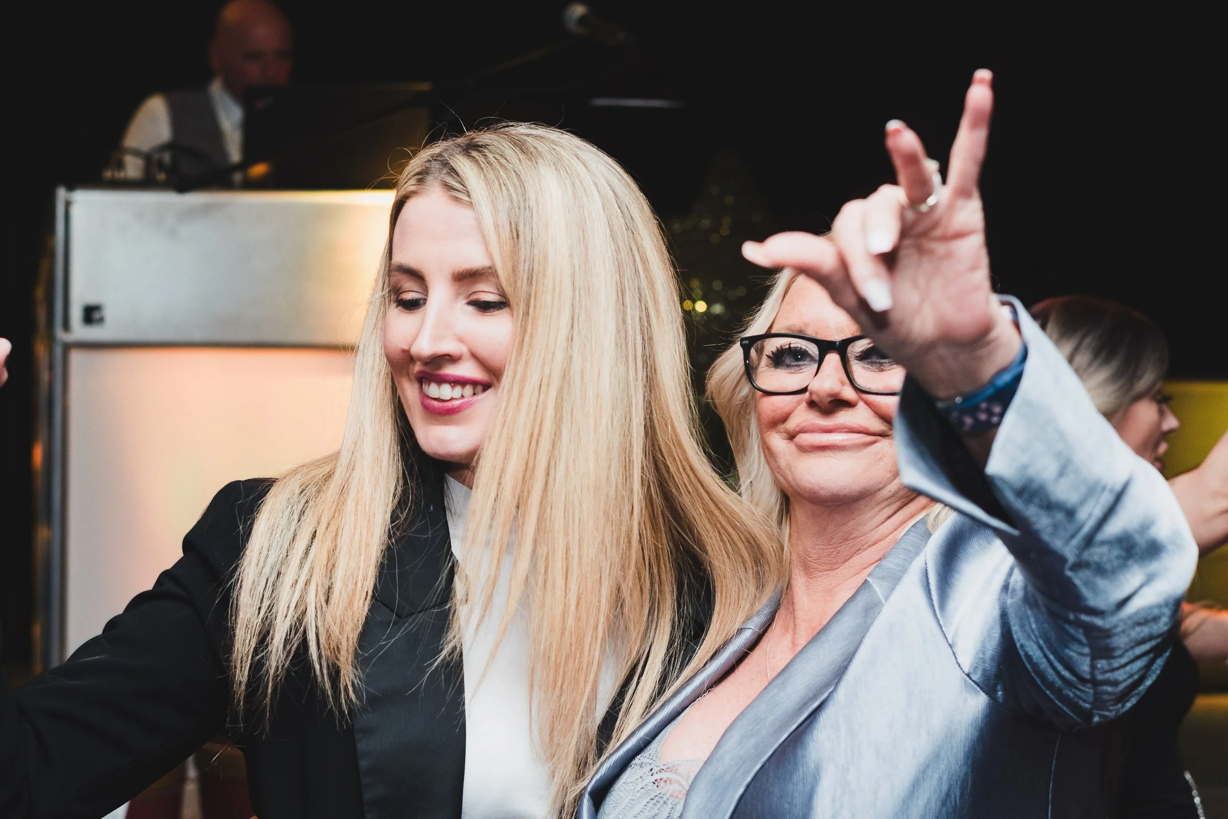 Two women taking a selfie together at an event. One woman with long blonde hair and a big smile, the other woman with short blonde hair and glasses, making a peace sign with her hand.