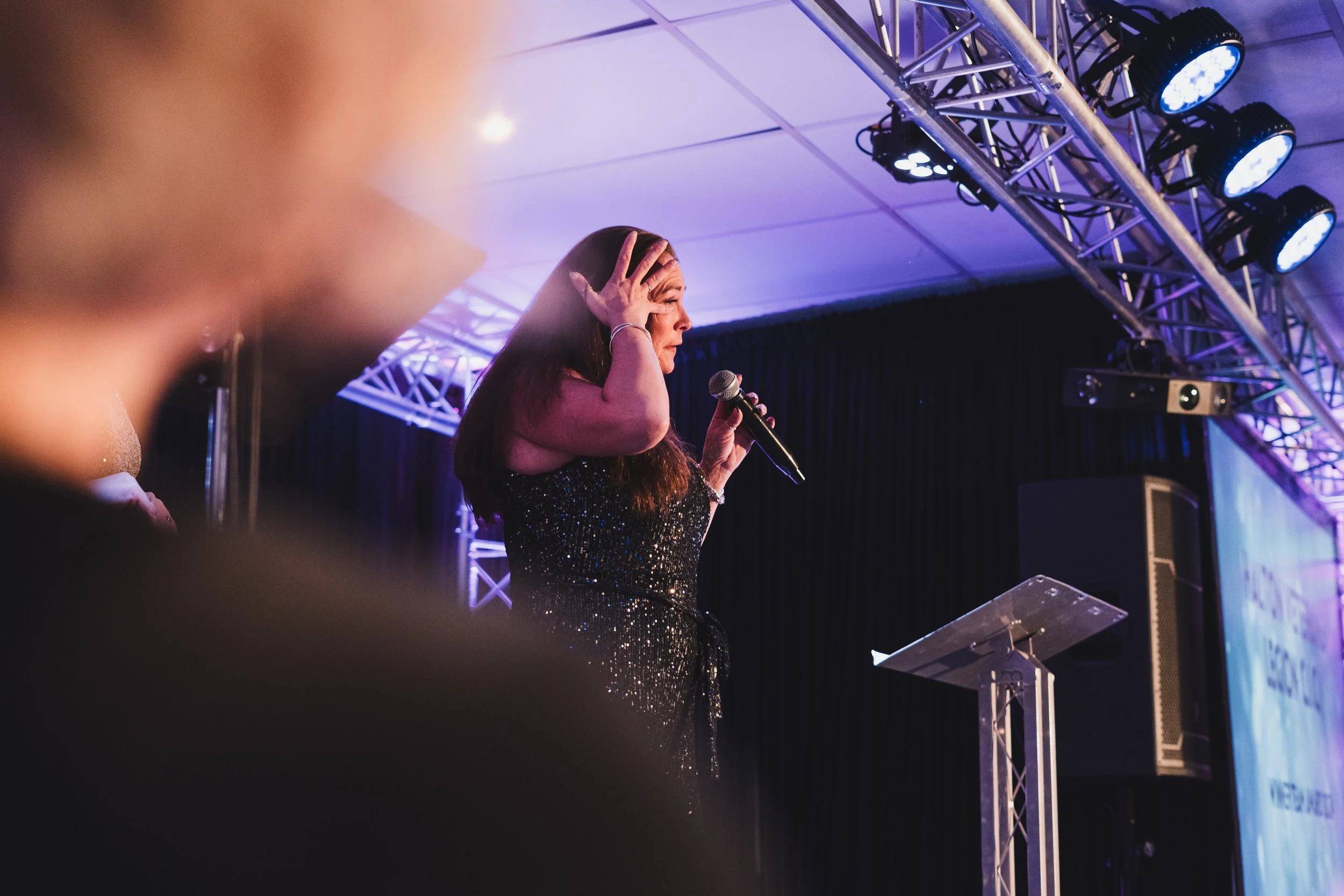 A woman in a sparkly black dress holding a microphone on stage with stage lights overhead.