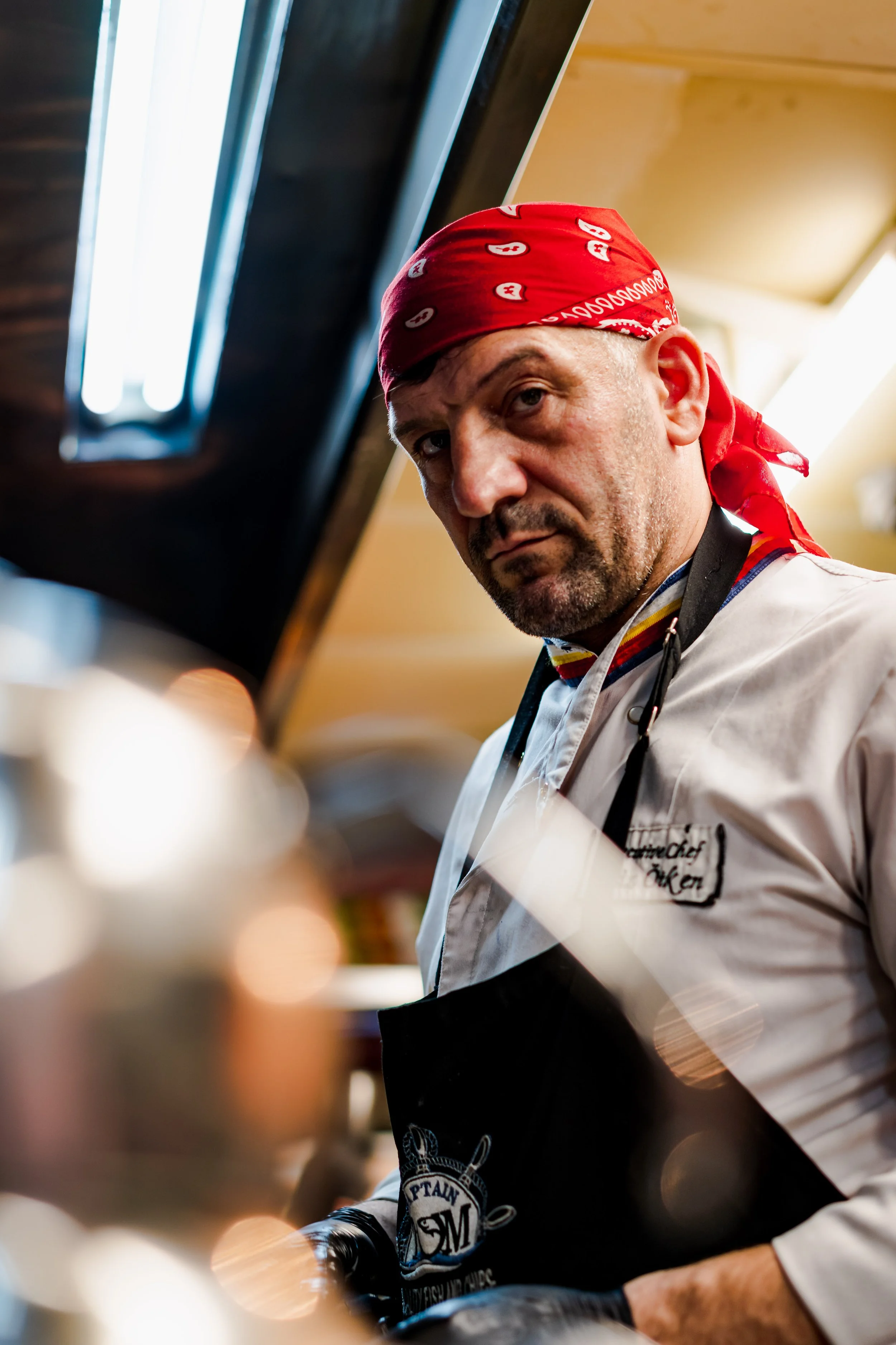 A middle-aged man wearing a red bandana, chef's coat, and black apron looking into the camera in a kitchen environment.