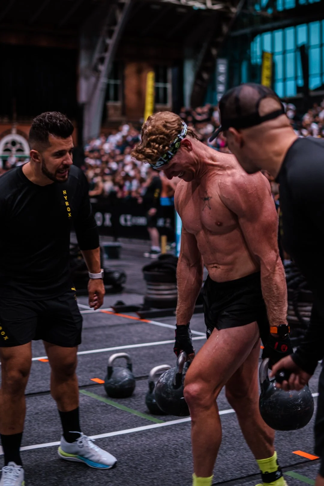 A shirtless man with a muscular build and curly hair is lifting kettlebells while surrounded by two other men in workout gear during a fitness event in a gym with a high arched ceiling and an audience in the background.
