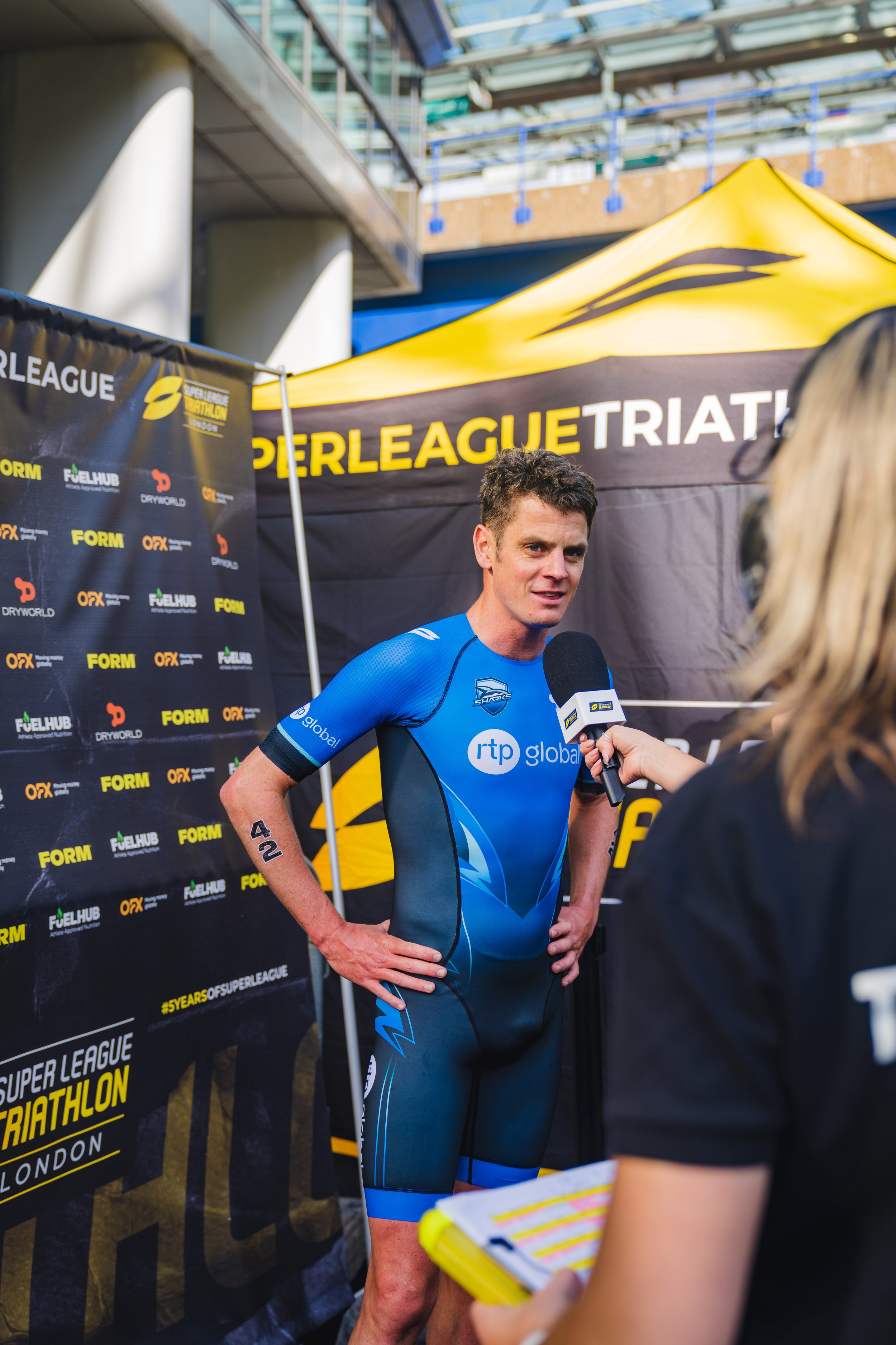 A man in a blue triathlon suit is being interviewed by a woman holding a microphone and notepad. They are standing in front of a black and yellow banner at a Super League Triathlon event in London, with a yellow tent overhead.