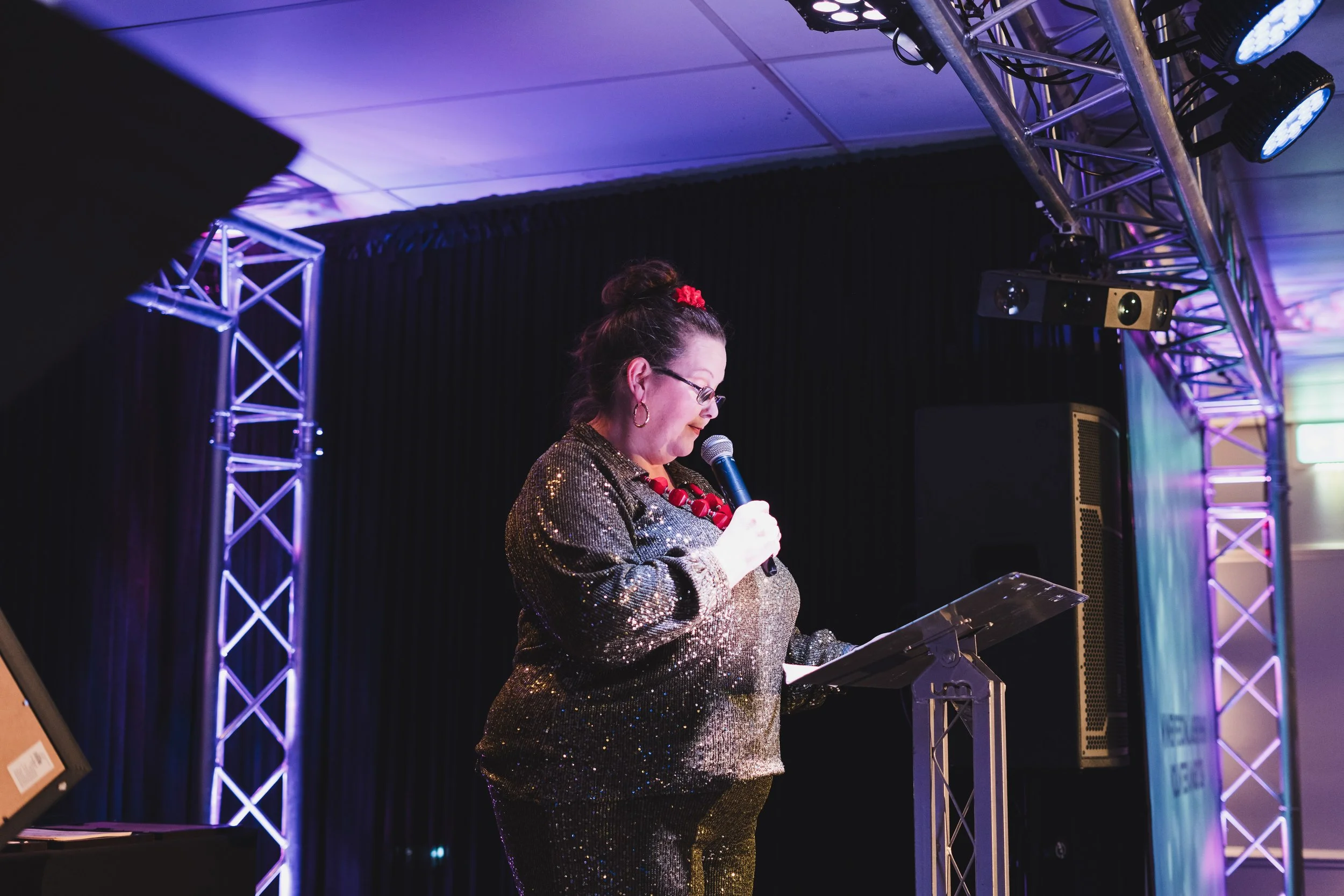 A woman in a gold, glittery outfit speaking into a microphone at a podium on a stage with truss structure and lighting.