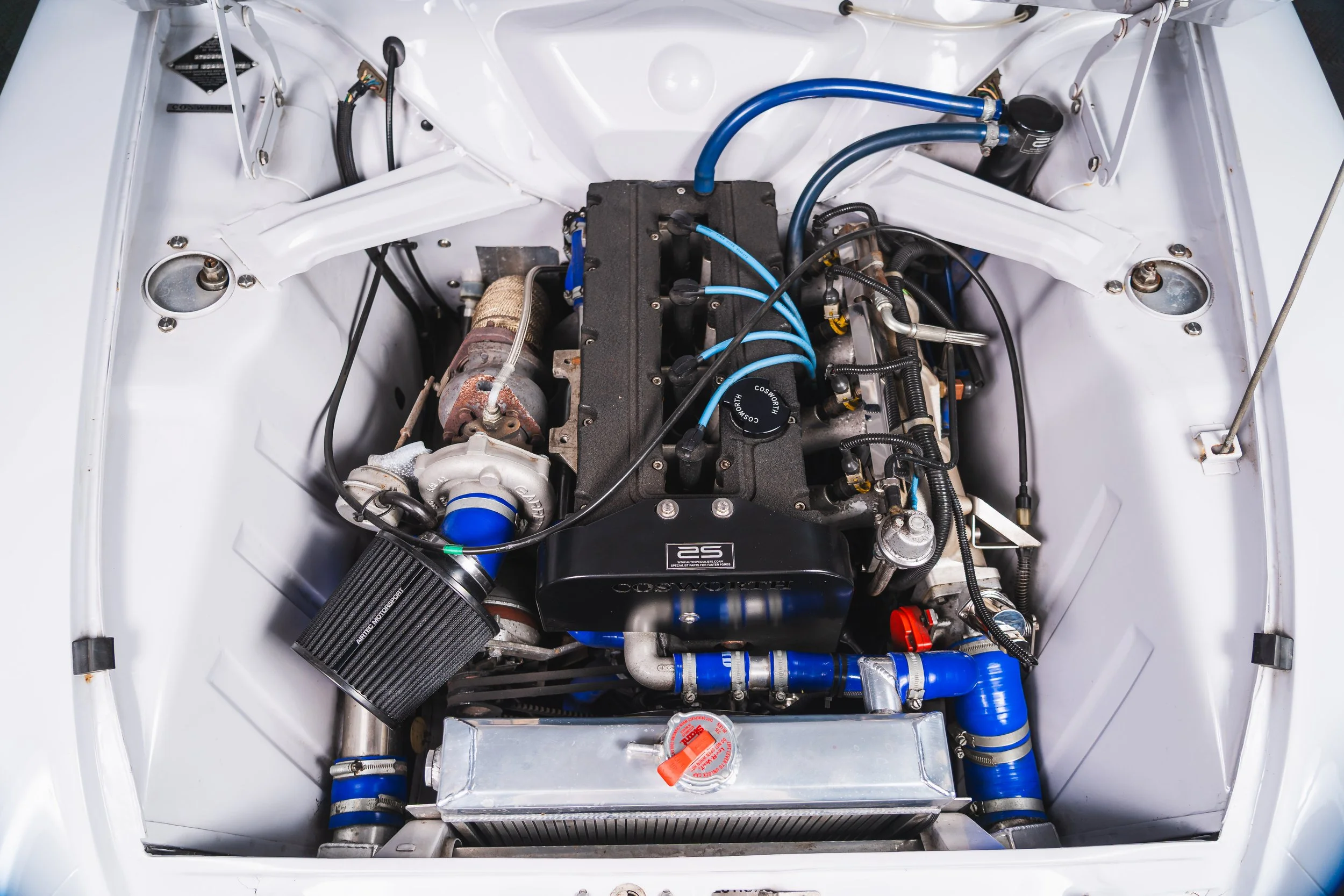 View of a car engine in a white engine bay with various components, hoses, and wiring.