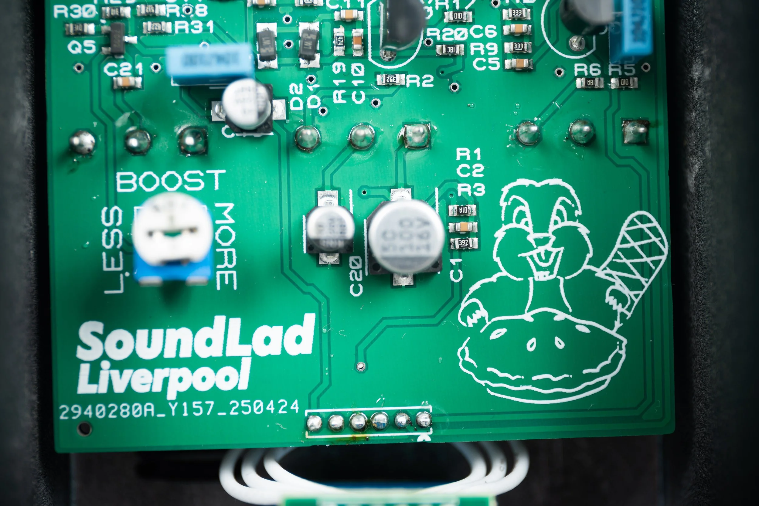 Close-up photo of a green printed circuit board from SoundLad Liverpool with electronic components and a white illustration of a smiling cartoon character with a hammer, holding a pie.
