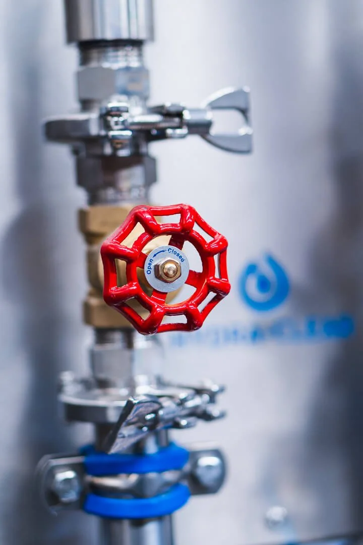 Close-up of a red valve handle connected to a metallic pipe, with a white circular label indicating 'Open - Close', in a mechanical or industrial setting.