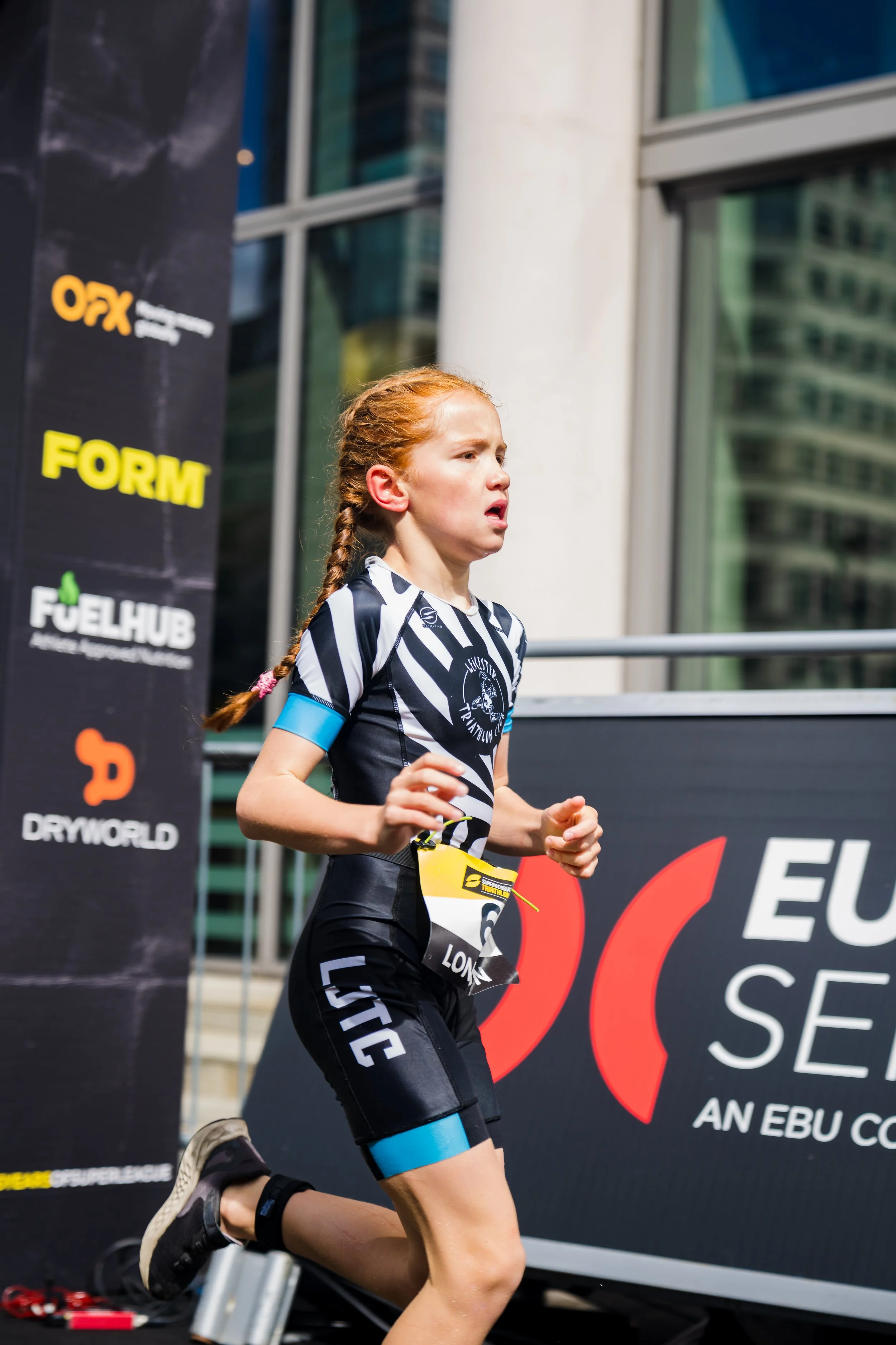 A young girl with red hair running in a triathlon event, wearing a black and white striped athletic outfit and a blue wristband, with a focused expression.