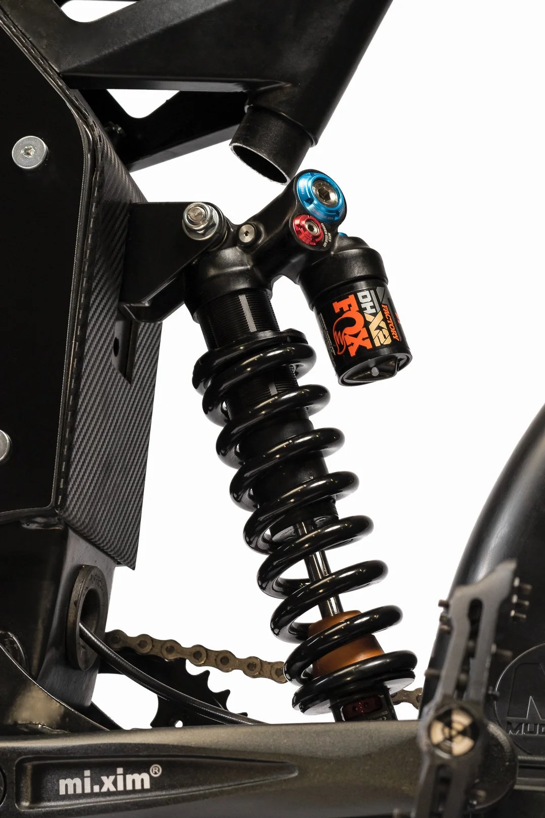 Close-up of a mountain bike's rear suspension system, featuring a coil shock absorber with a Fox logo, mounted on a black frame with carbon fiber elements.
