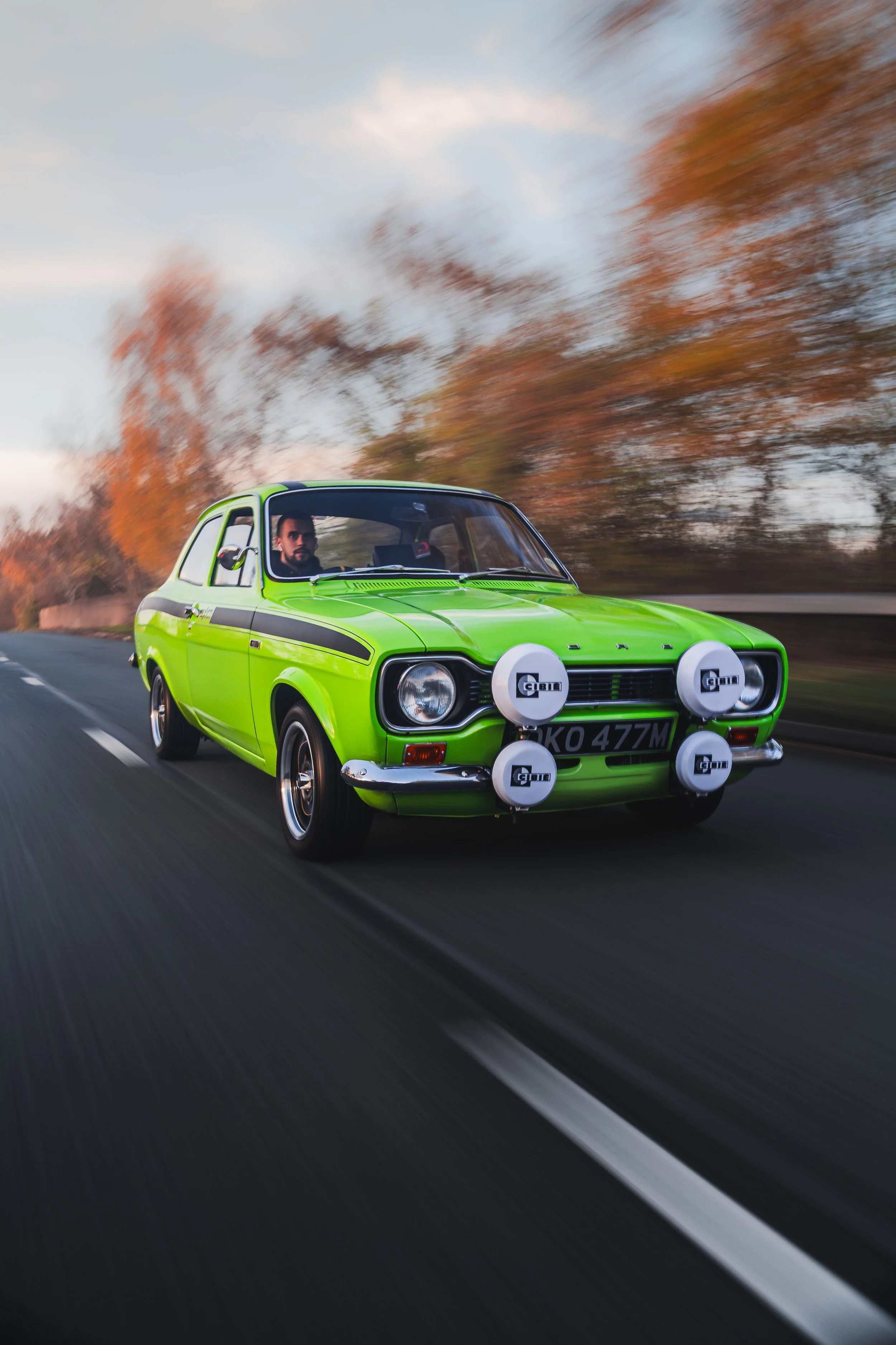 A vintage bright green Ford car with four rally lights mounted on the front, driving on a highway surrounded by autumn trees, with a person visible inside.