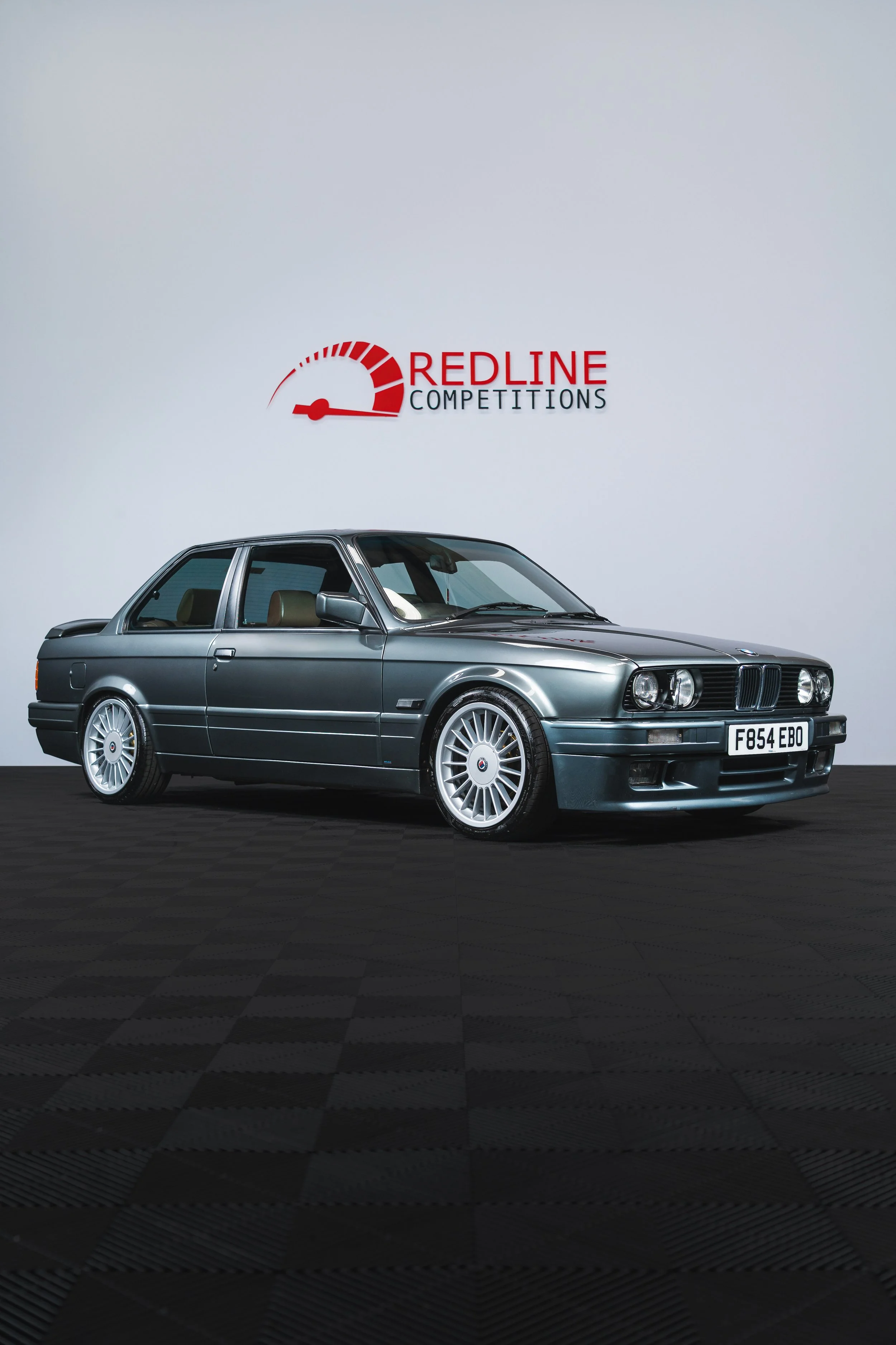 A classic BMW E30 car in metallic gray color, displayed at an auction or showroom, with a 'Redline Competitions' sign in the background.