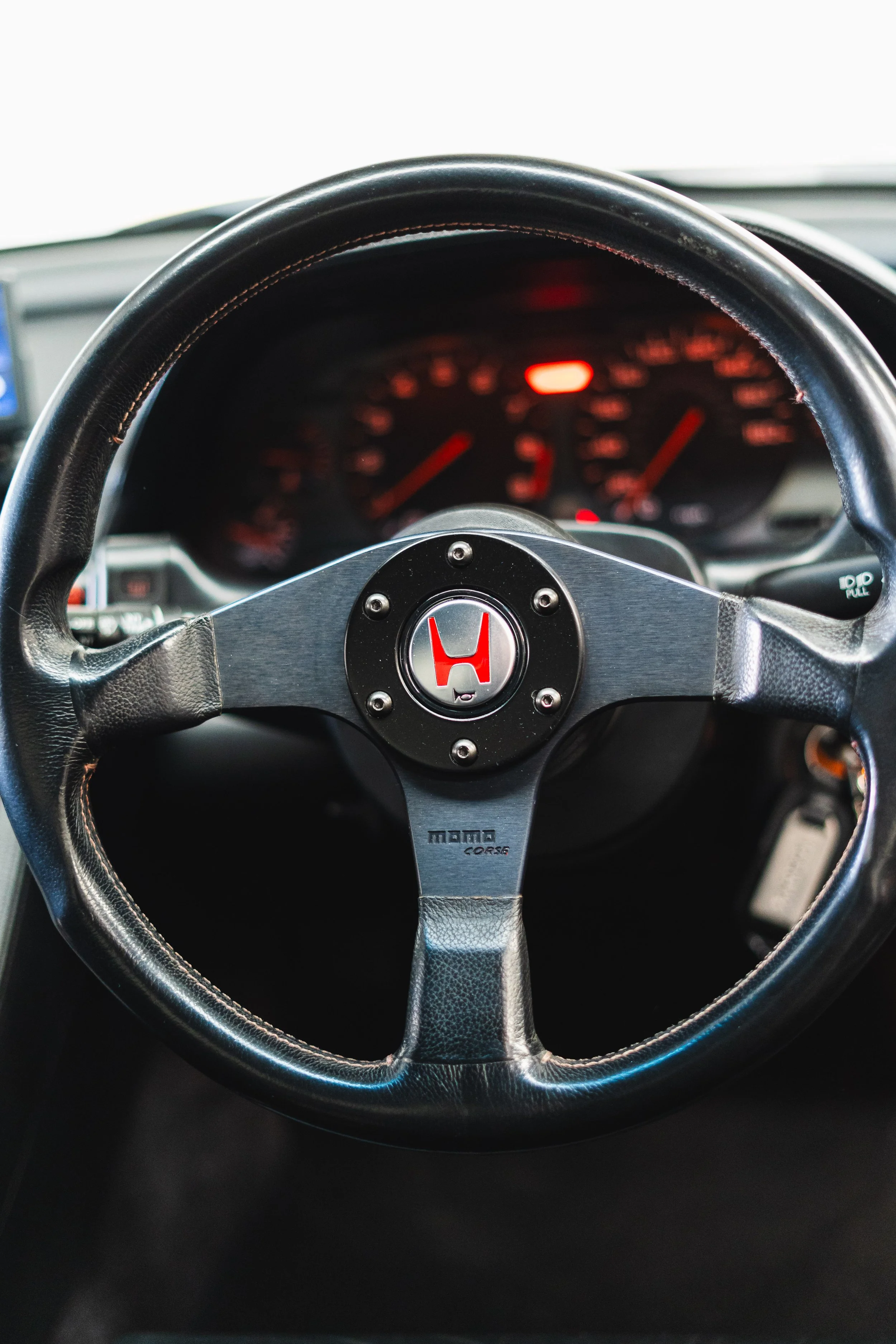 Close-up of a black Momo Corse steering wheel with a silver center and red emblem, inside a car dashboard showing speed and RPM gauges.