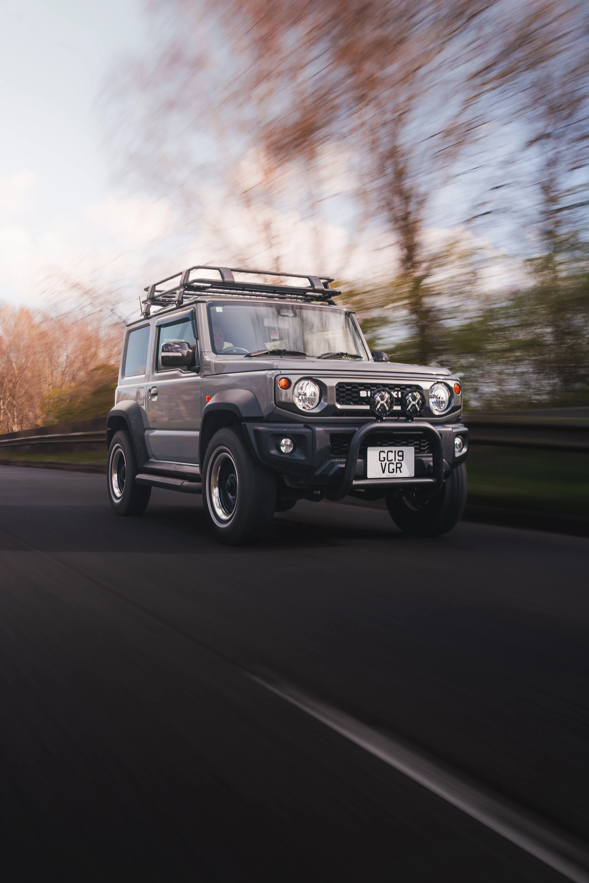A gray Suzuki Jimny driving on a road with autumn trees in the background.