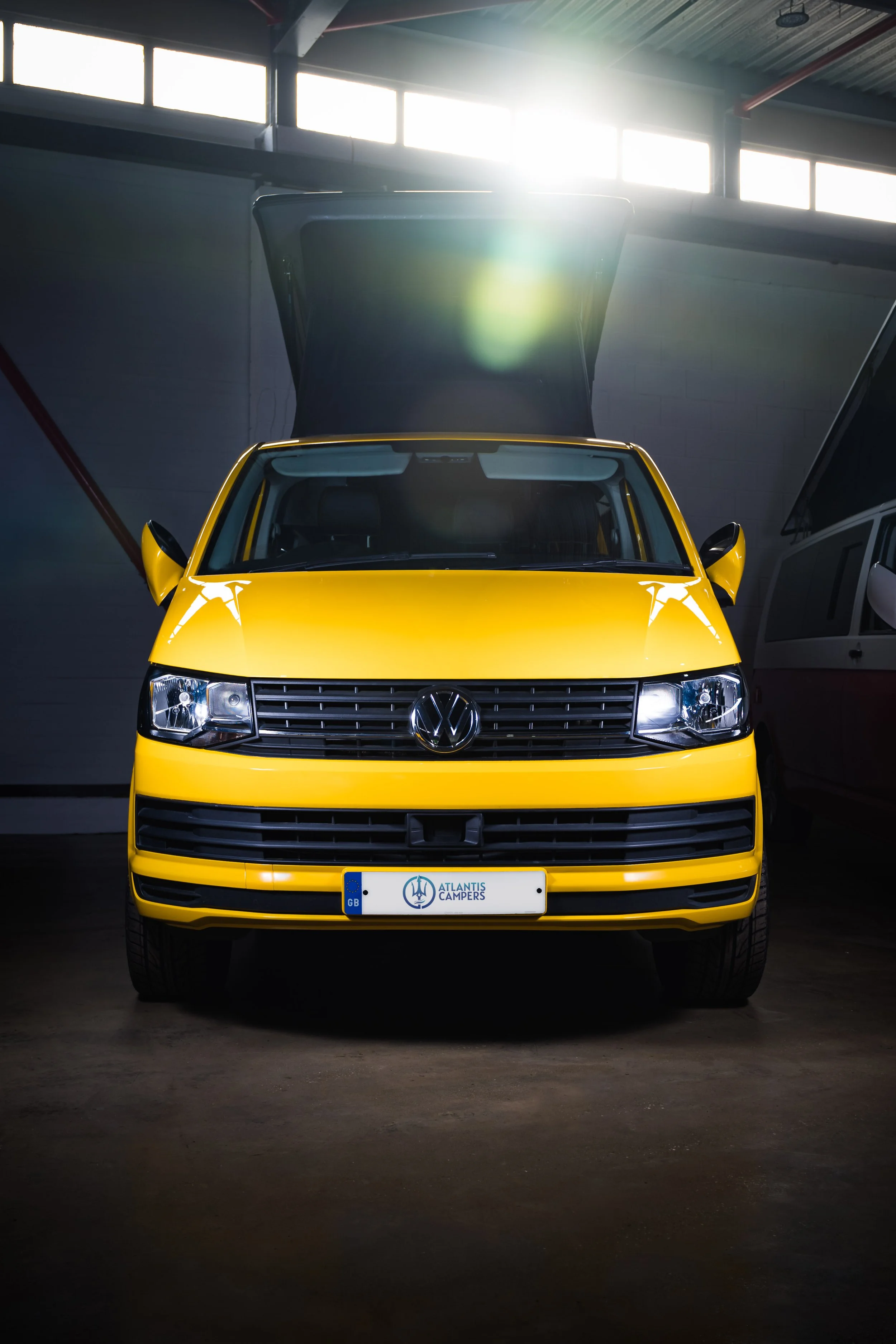 A yellow Volkswagen camper van parked inside a building with the front facing forward. The van has a Volkswagen emblem on the grille, and a license plate reading 'ATLAS CAMPERS' from the UK. The van's roof is partially opened, and sunlight is streami