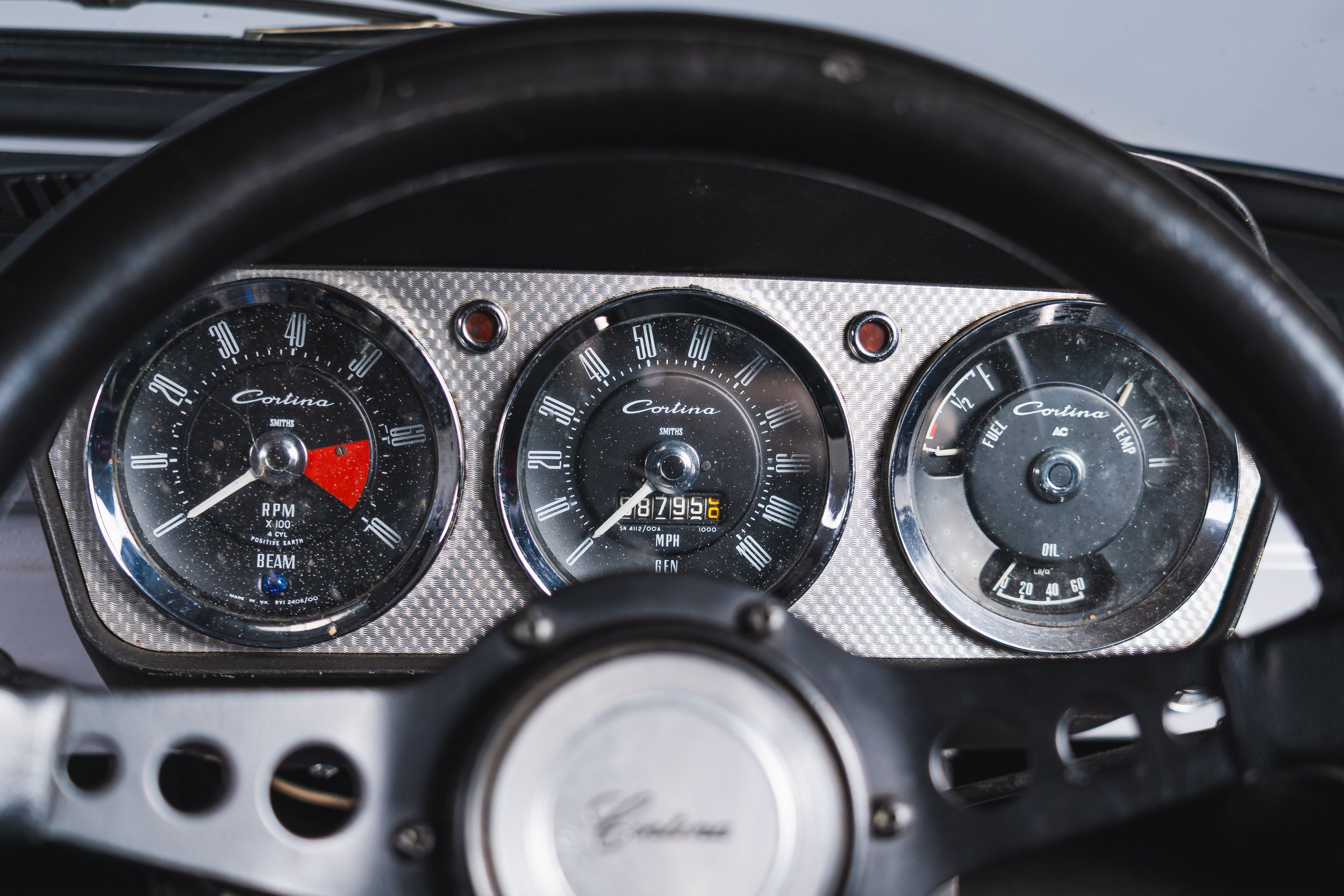 The dashboard of a vintage car showing three round gauges: left gauge displaying RPM with a red zone, center gauge showing speed up to 120 MPH, right gauge with fuel, oil, temperature, and voltage indicators. The steering wheel is partly visible at t