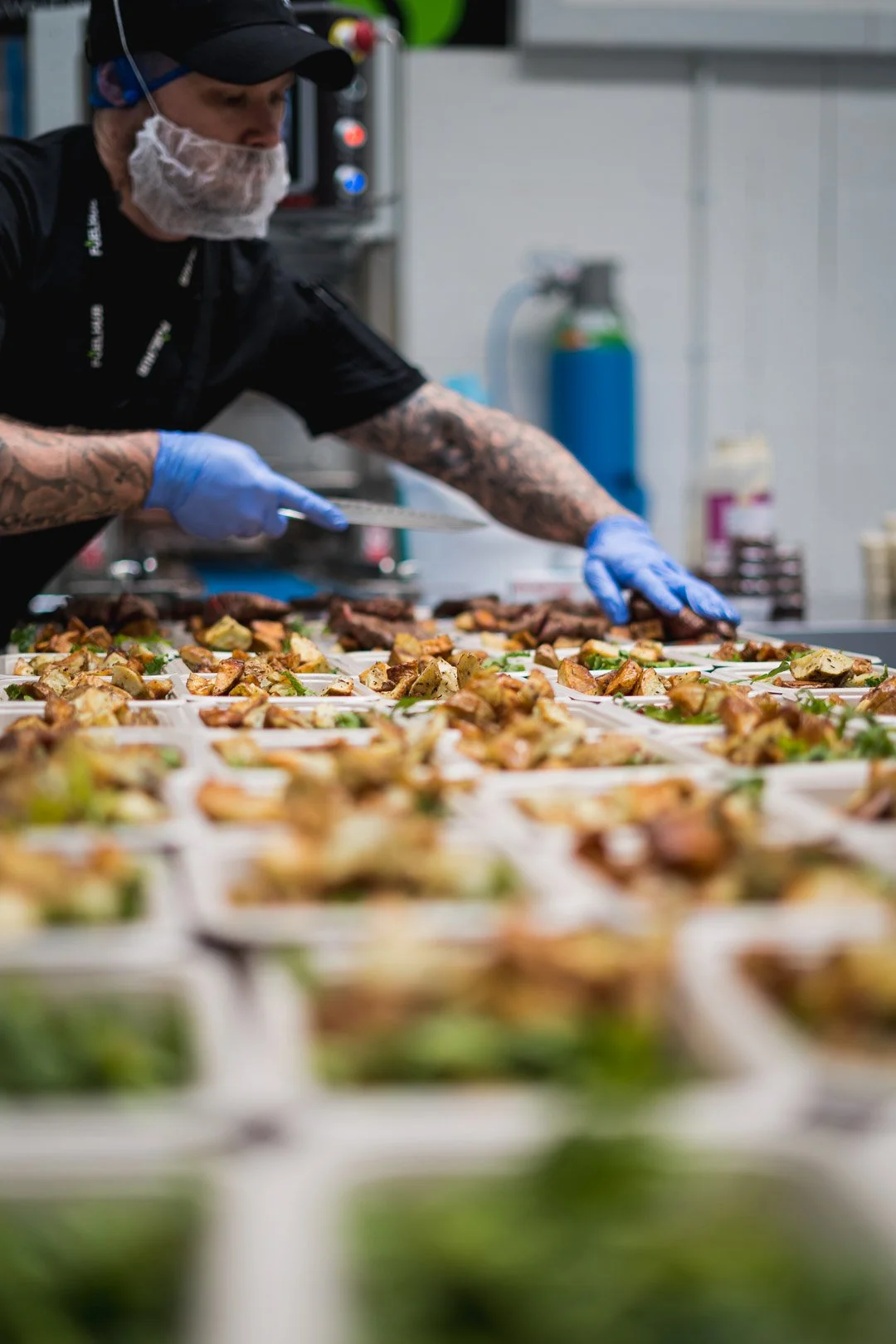 A chef with tattoos wearing a black cap, black shirt, blue gloves, and a hairnet preparing multiple food trays in a professional kitchen.