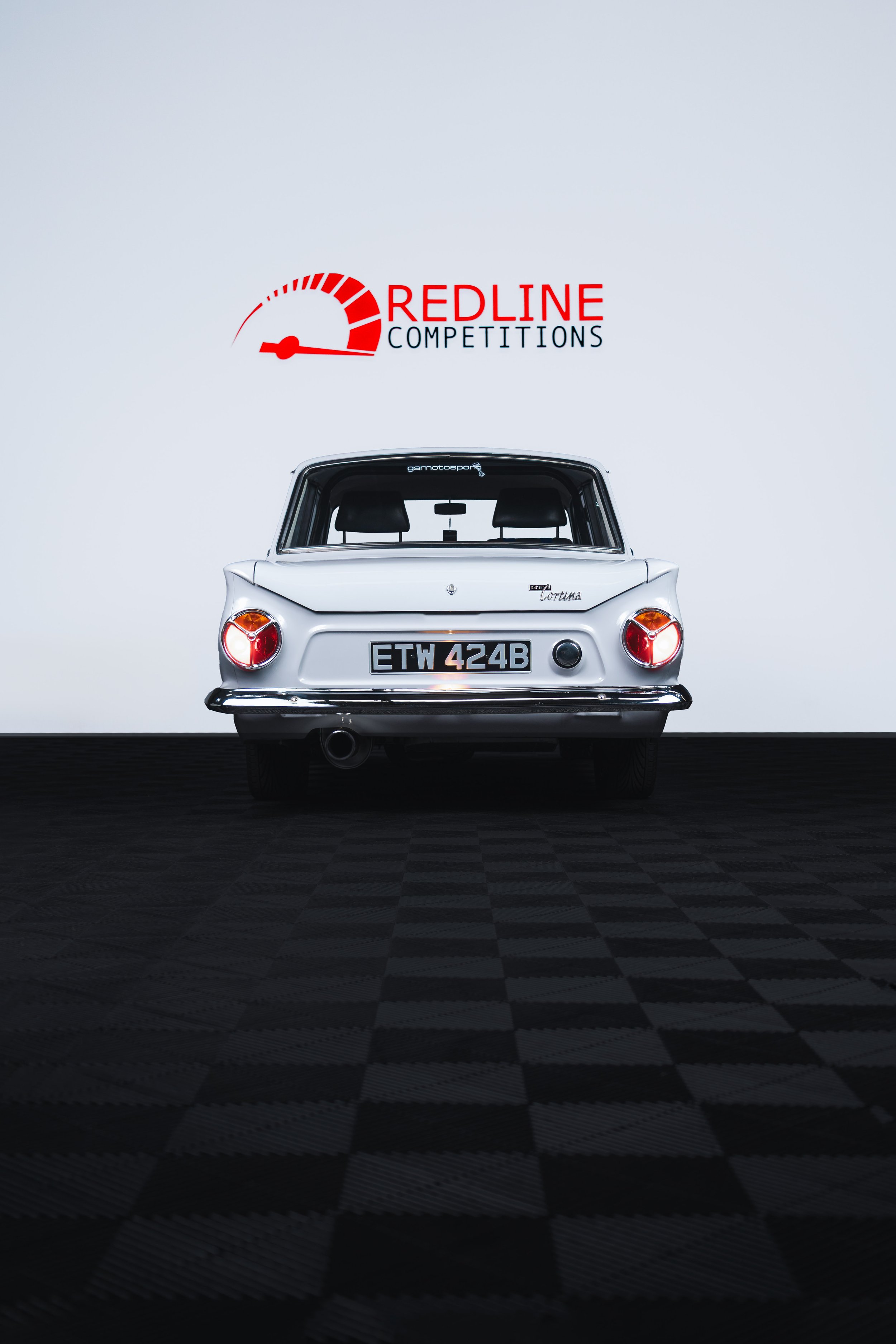 Rear view of a classic white car with black and red license plate, parked on a checkered floor in front of a white wall with ‘Redline Competitions’ logo and a speedometer graphic.